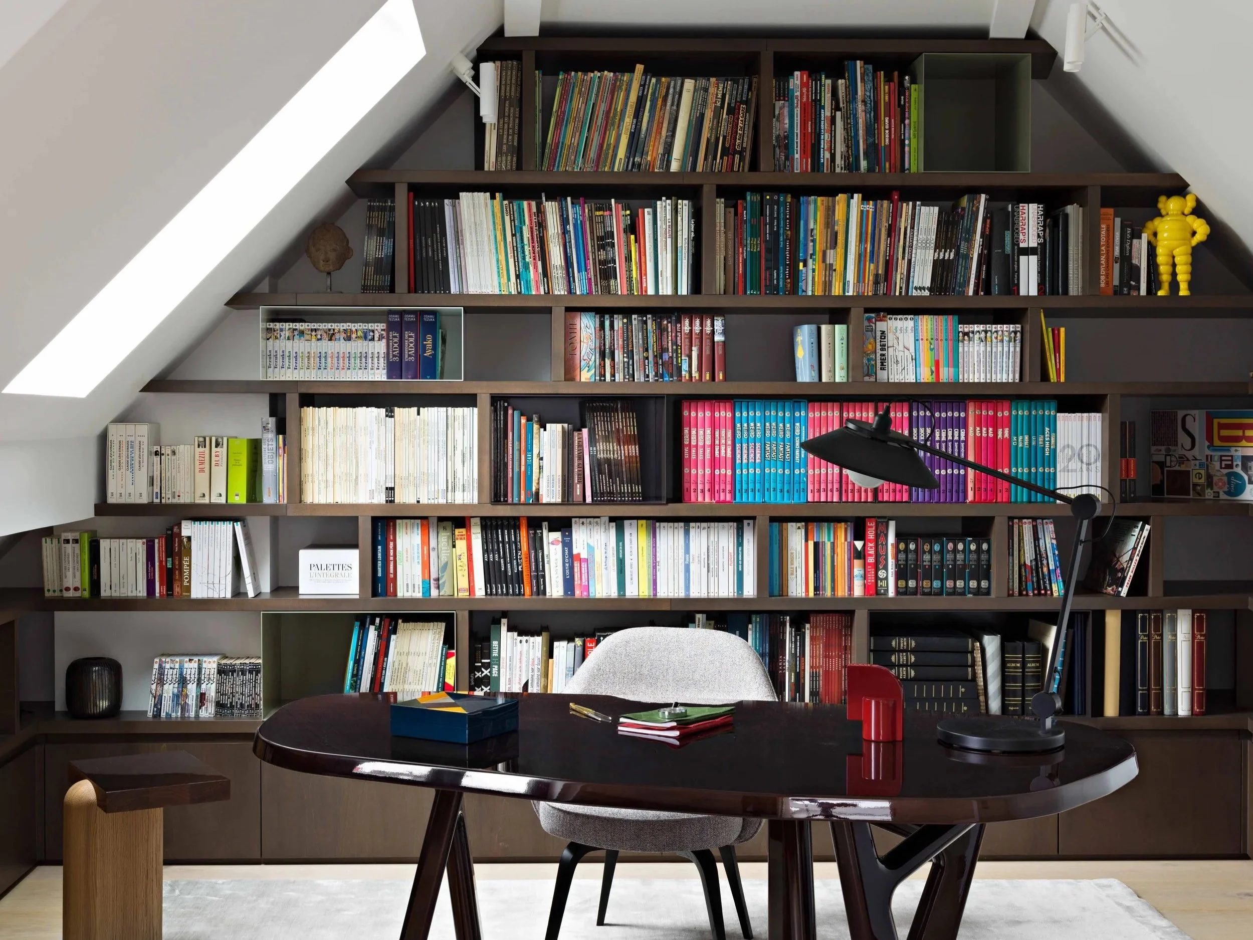 Une bibliothèque en forme de triangle remplie de livres colorés, avec un bureau et une chaise en premier plan, une lampe de bureau noire, et quelques objets décoratifs jaunes et noirs.