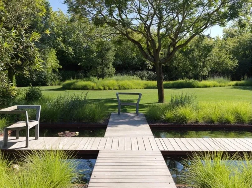 Jardin calme avec une petite écluse en bois, deux bancs en métal et un arbre au centre, entouré de verdure.