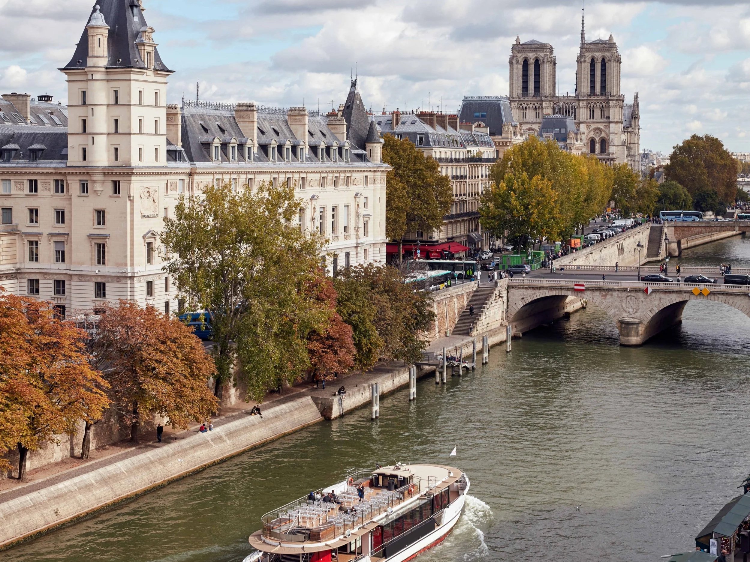 Vue de la Seine à Paris avec un bateau naviguant, des arbres aux couleurs d'automne, un pont en pierre, et la cathédrale Notre-Dame en arrière-plan.