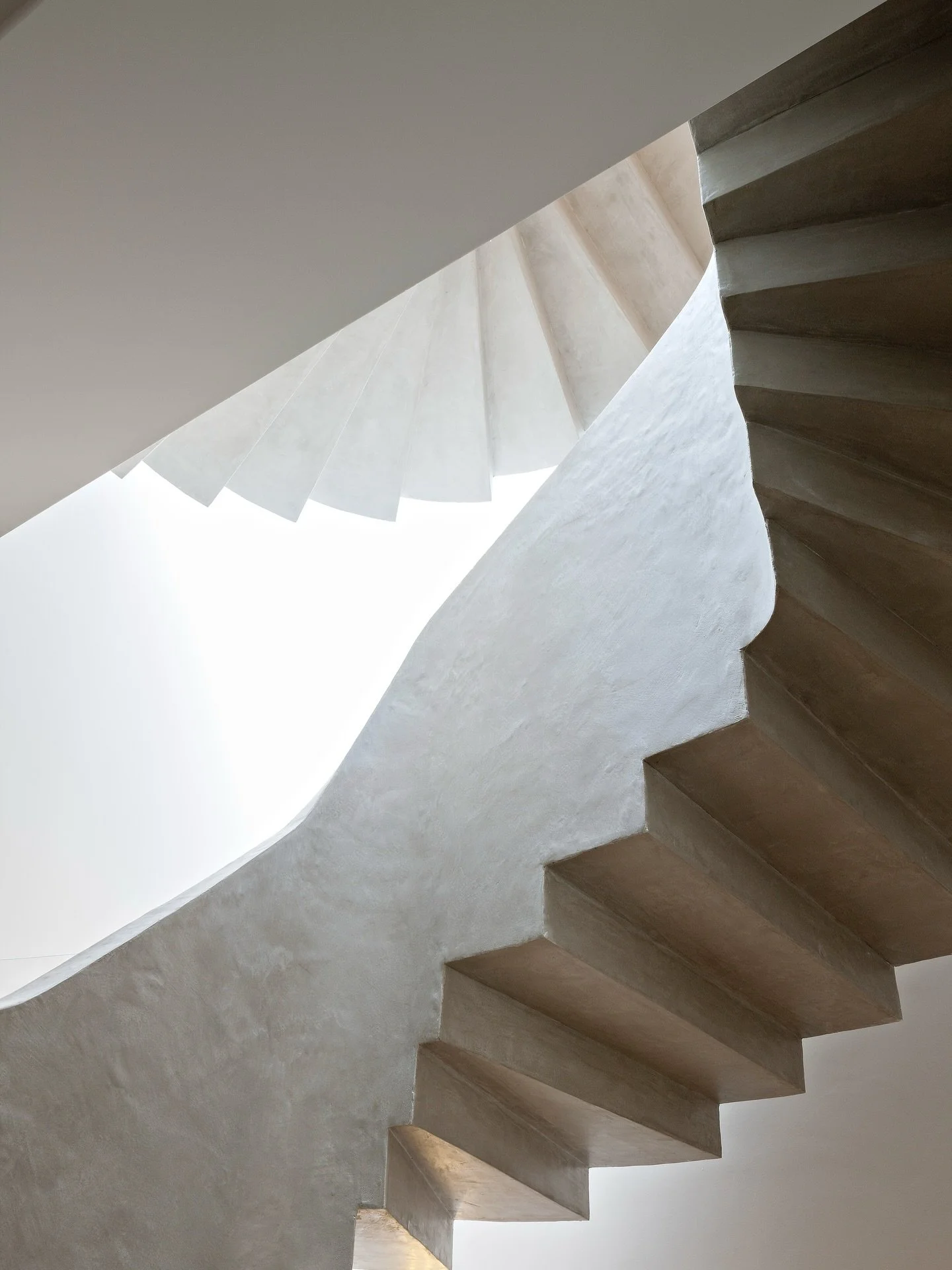 A sweeping staircase carved in subtle curves, bathed in gentle daylight. The raw texture of the material meets the disciplined geometry of the steps, each angle, each shadow is quietly intentional. A moment of architecture where form, light and mater