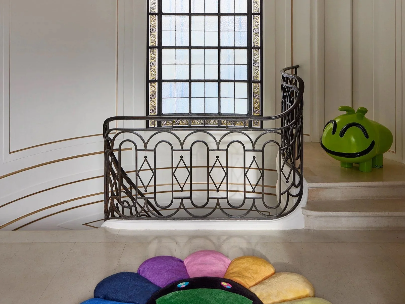 Intérieur d'une maison avec une rampe en fer noire, une fenêtre avec vitraux, un sol en pierre claire, une sculpture de couleur verte représentant un visage souriant, et un tapis coloré en forme de fleur en premier plan.