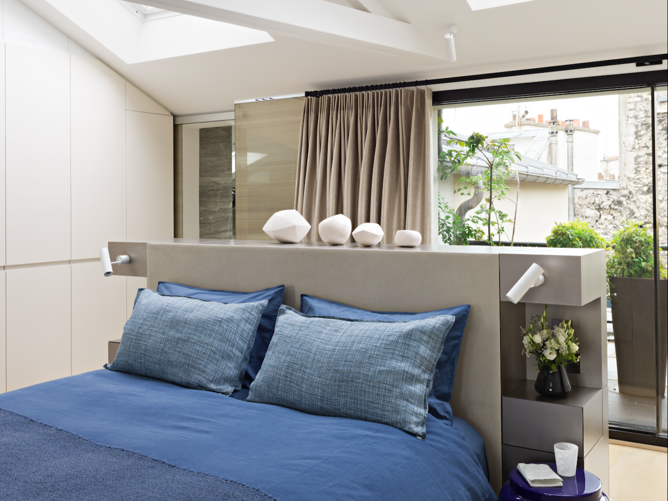 Chambre moderne avec un lit en bois clair avec des oreillers bleus, grande fenêtre avec vue sur un balcon et des plantes, décoration minimaliste avec vases blancs sur le mur au-dessus du lit.