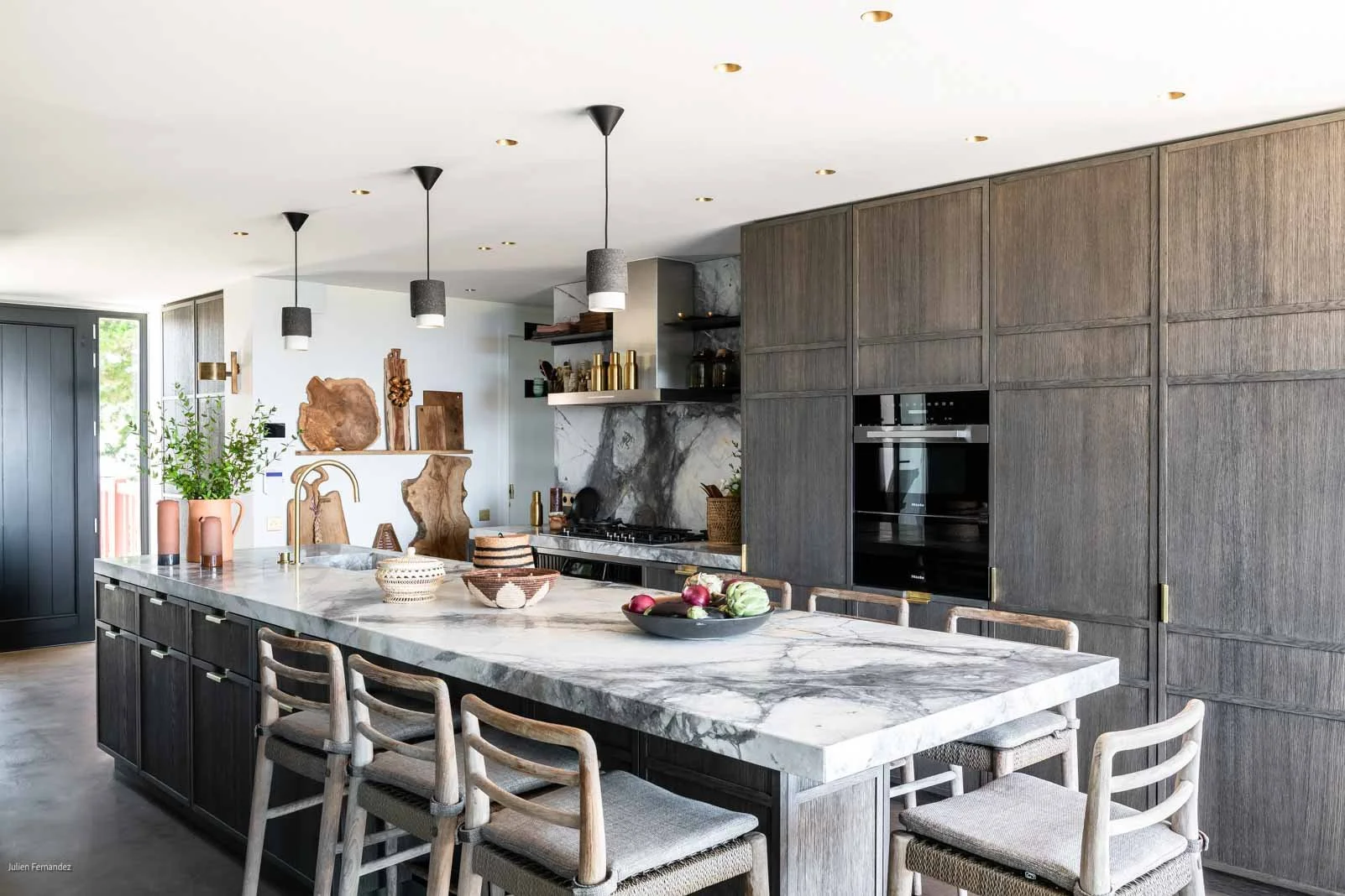 Cuisine moderne avec un grand plan de travail en marbre, armoires en bois gris foncé, îlot central, éclairage suspendu et décorations naturelles.