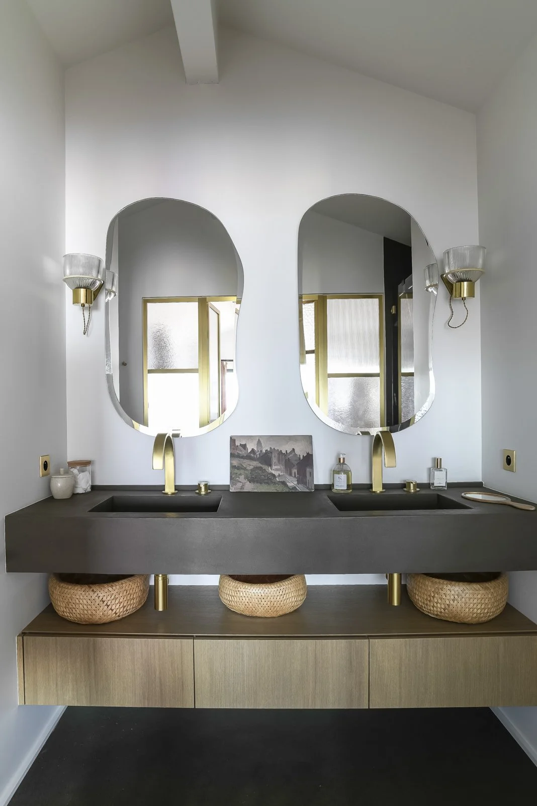 Une salle de bain moderne avec un double lavabo noir, deux miroirs ovales, deux appliques murales, un meuble en bois clair, et des paniers en osier sous le lavabo.