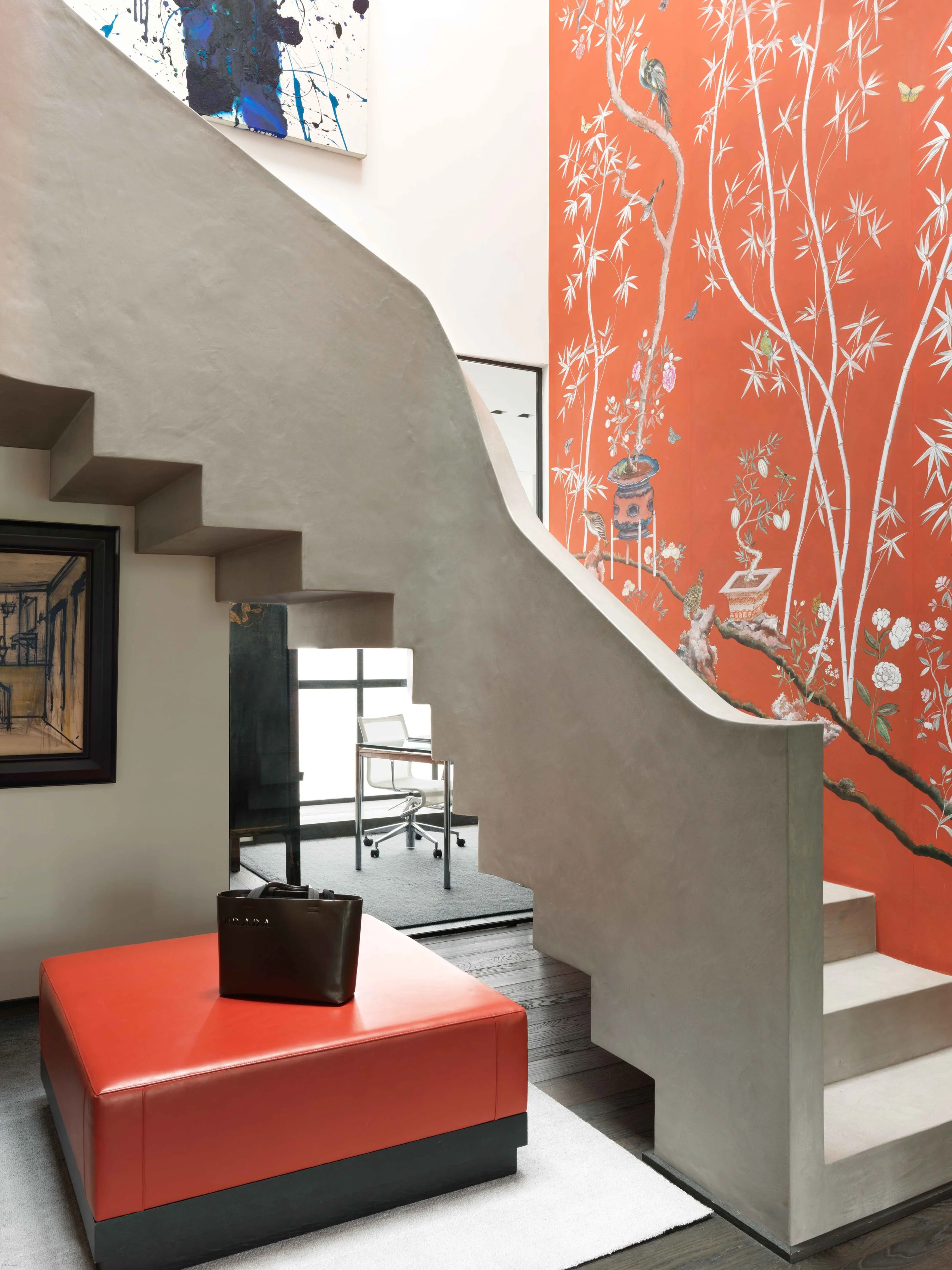 Intérieur moderne avec escalier en béton, mur peint avec un grand motif d'arbres et de oiseaux, meuble en cuir rouge, tableau sur le mur et un espace de bureau au fond avec une chaise de bureau.