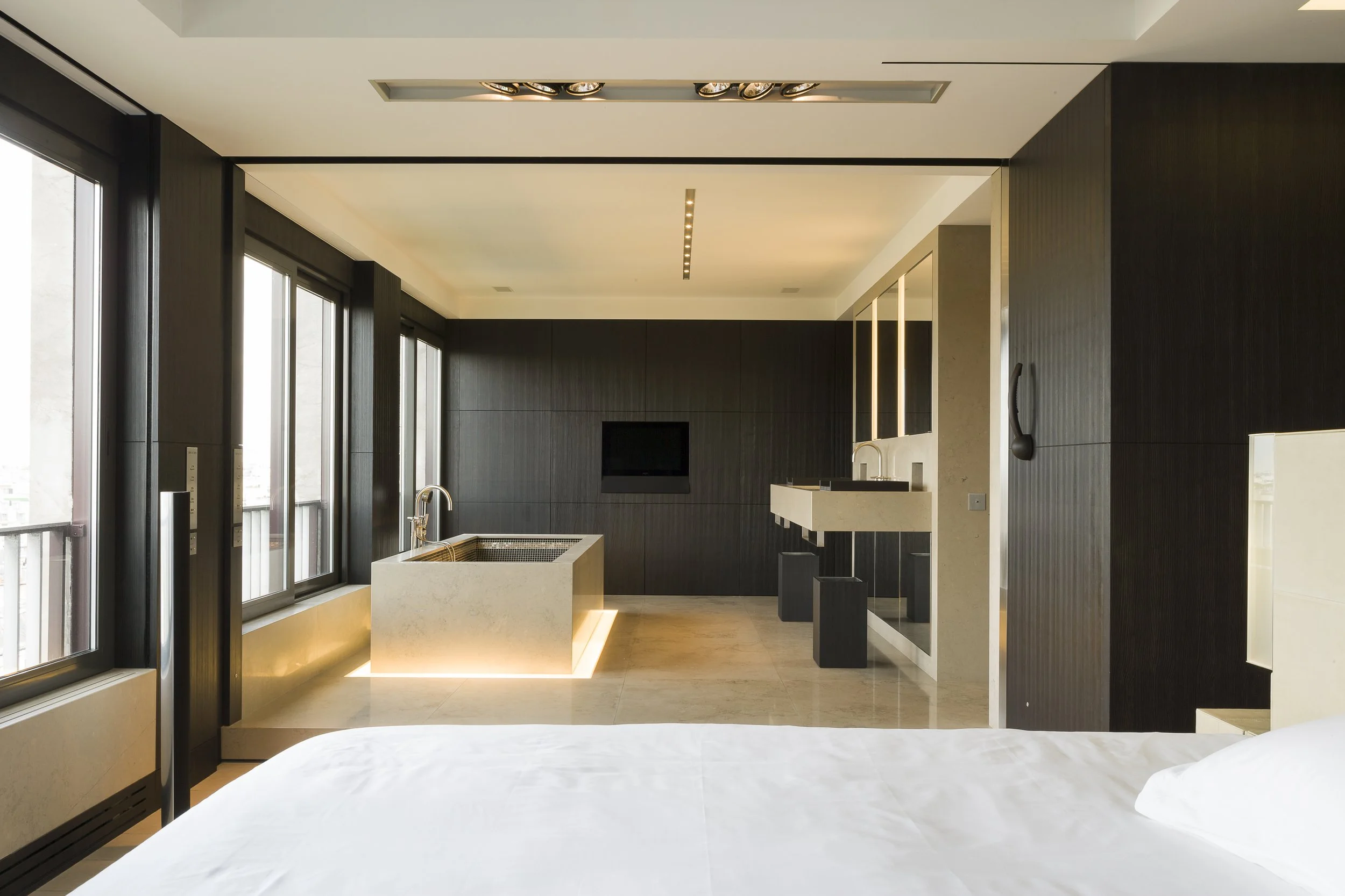 Intérieur moderne d'une chambre d'hôtel avec mur en bois foncé, grande fenêtre, lit blanc en premier plan, coin spa avec baignoire, télévision murale et lavabo, éclairage intégré.