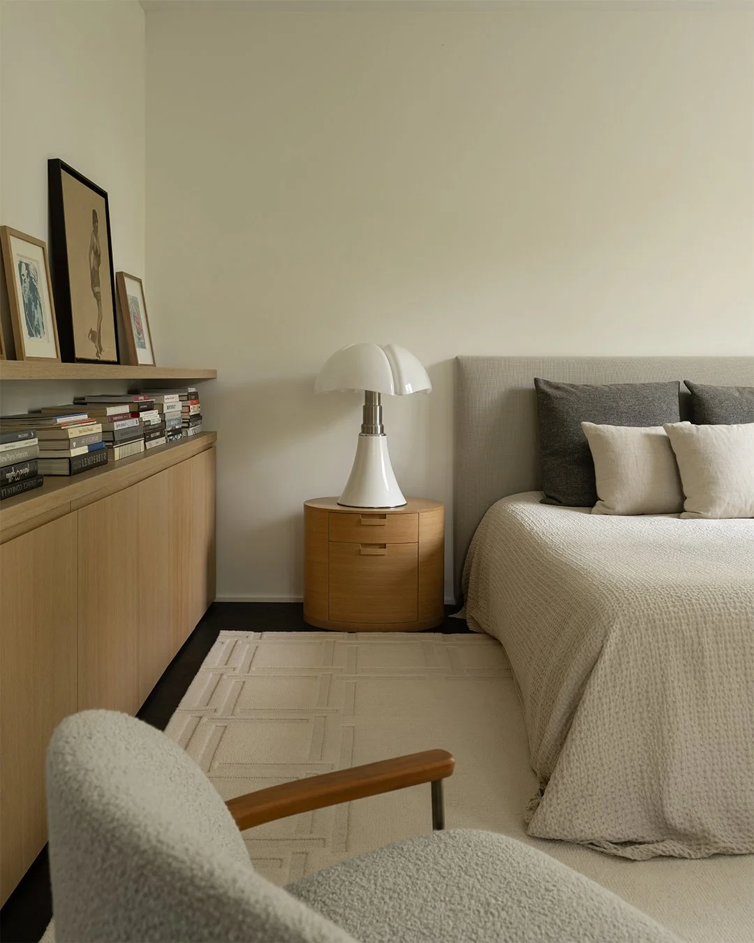 Coin de chambre avec lit king-size, table de nuit en bois avec lampe blanche, bibelots et cadres, fauteuil en tissu beige avec bras en bois, bibliothèque en bois clair avec livres et cadres d'art.