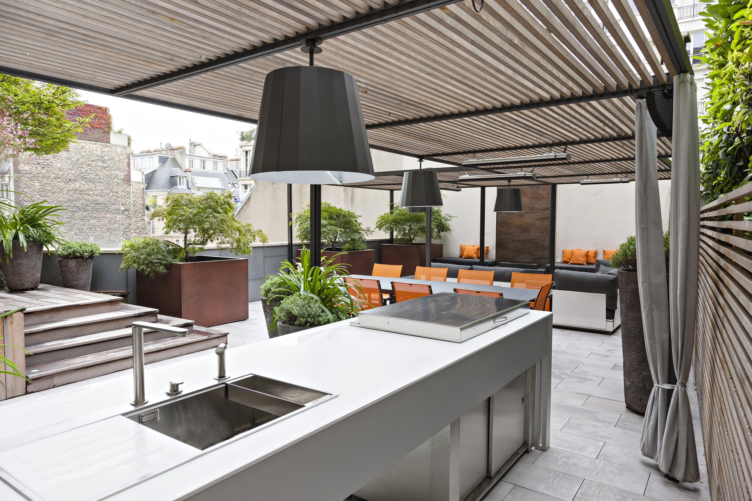 Terrasse urbaine avec mobilier confortable, plantes vertes, éclairage suspendu, et vue sur les bâtiments voisins.
