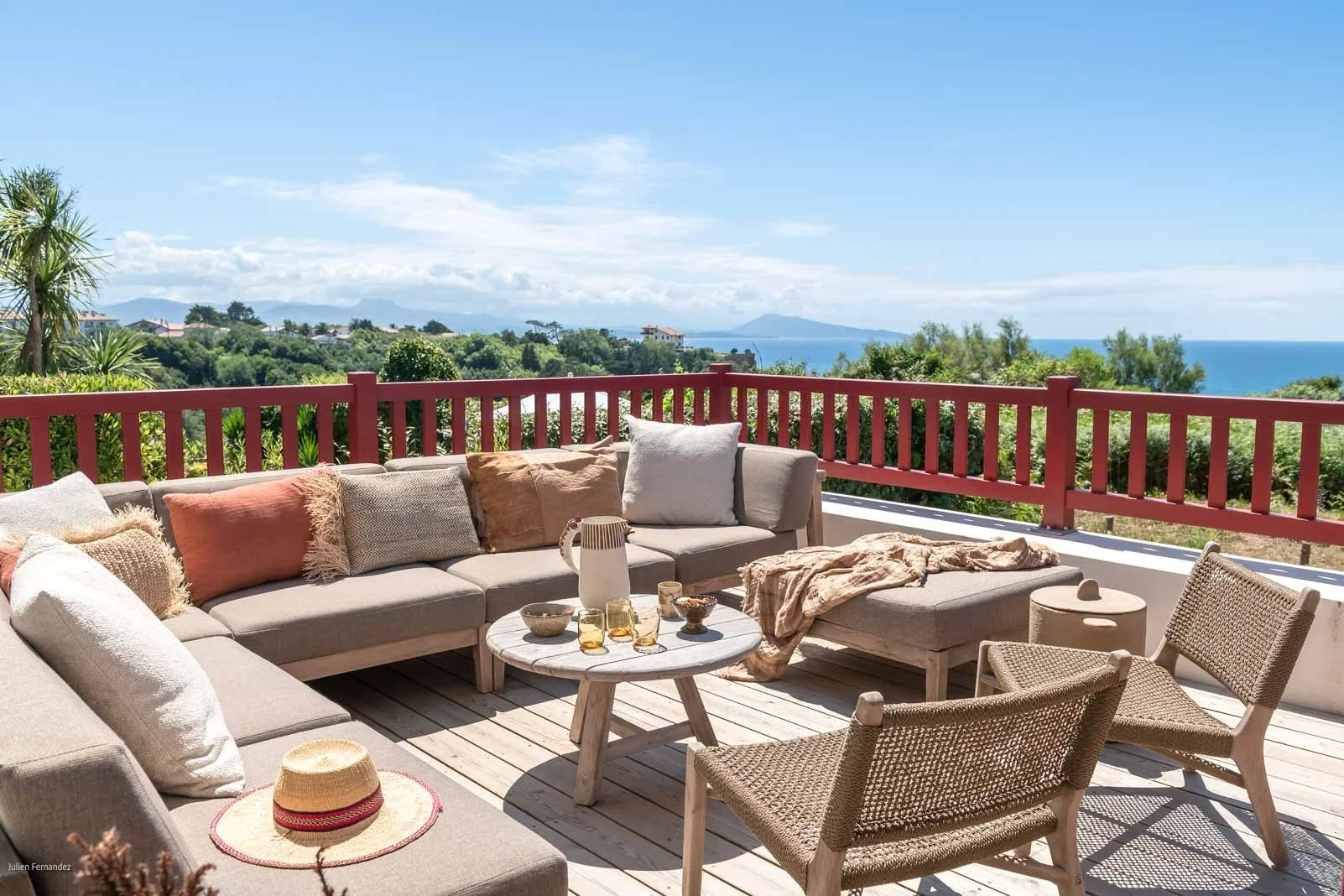 Terrasse avec canapé en bois et coussins, chaises en bois, vue sur la mer, ciel bleu, et végétation luxuriante.