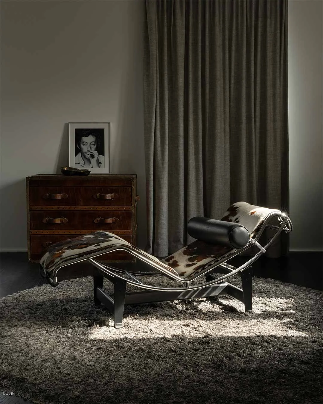 Une chaise longue design avec un motif en peau de vache, placée sur un tapis en laine, dans une pièce sombre avec une fenêtre à volets gris foncé, un meuble en bois vintage, une photo en noir et blanc d'une femme et des rideaux gris.