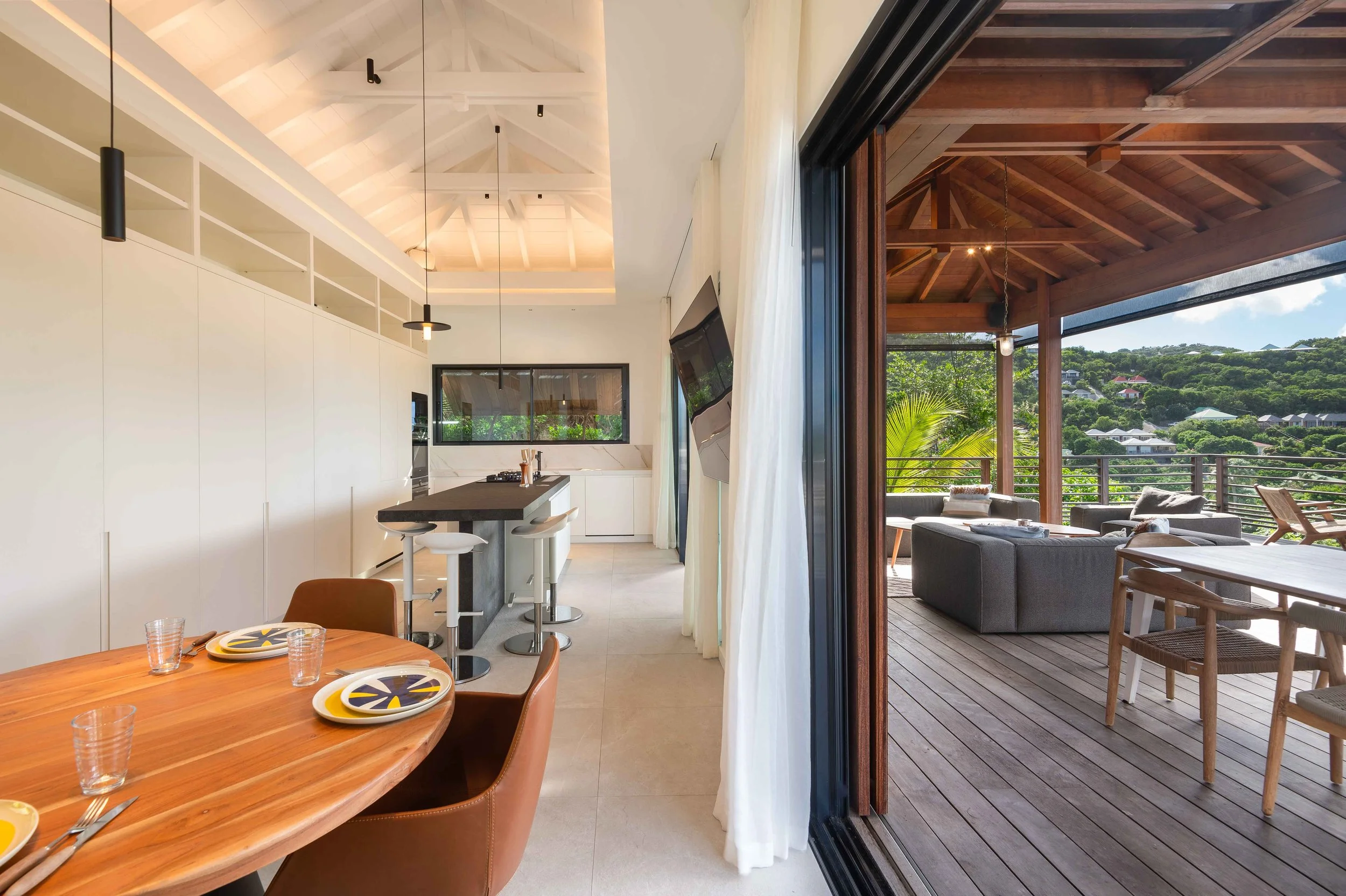Intérieur d'une maison moderne avec cuisine et salle à manger, vue sur un balcon en bois meublé avec des canapés et une vue panoramique sur la nature.