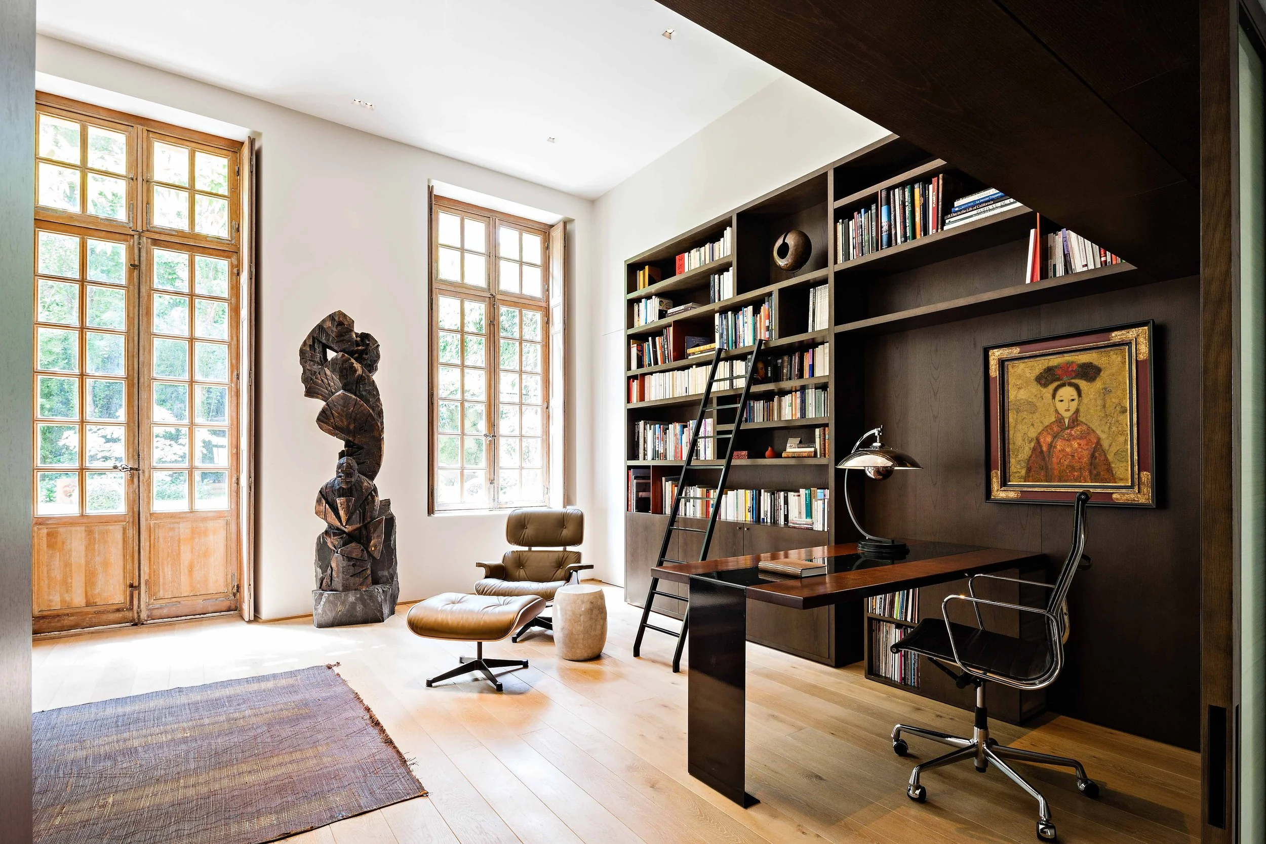 Un bureau avec deux grandes fenêtres en bois, une bibliothèque noire remplie de livres, un fauteuil en cuir avec repose-pieds, une sculpture en bois, un mur blanc, un tapis, un bureau en bois avec une lampe et une chaise de bureau.