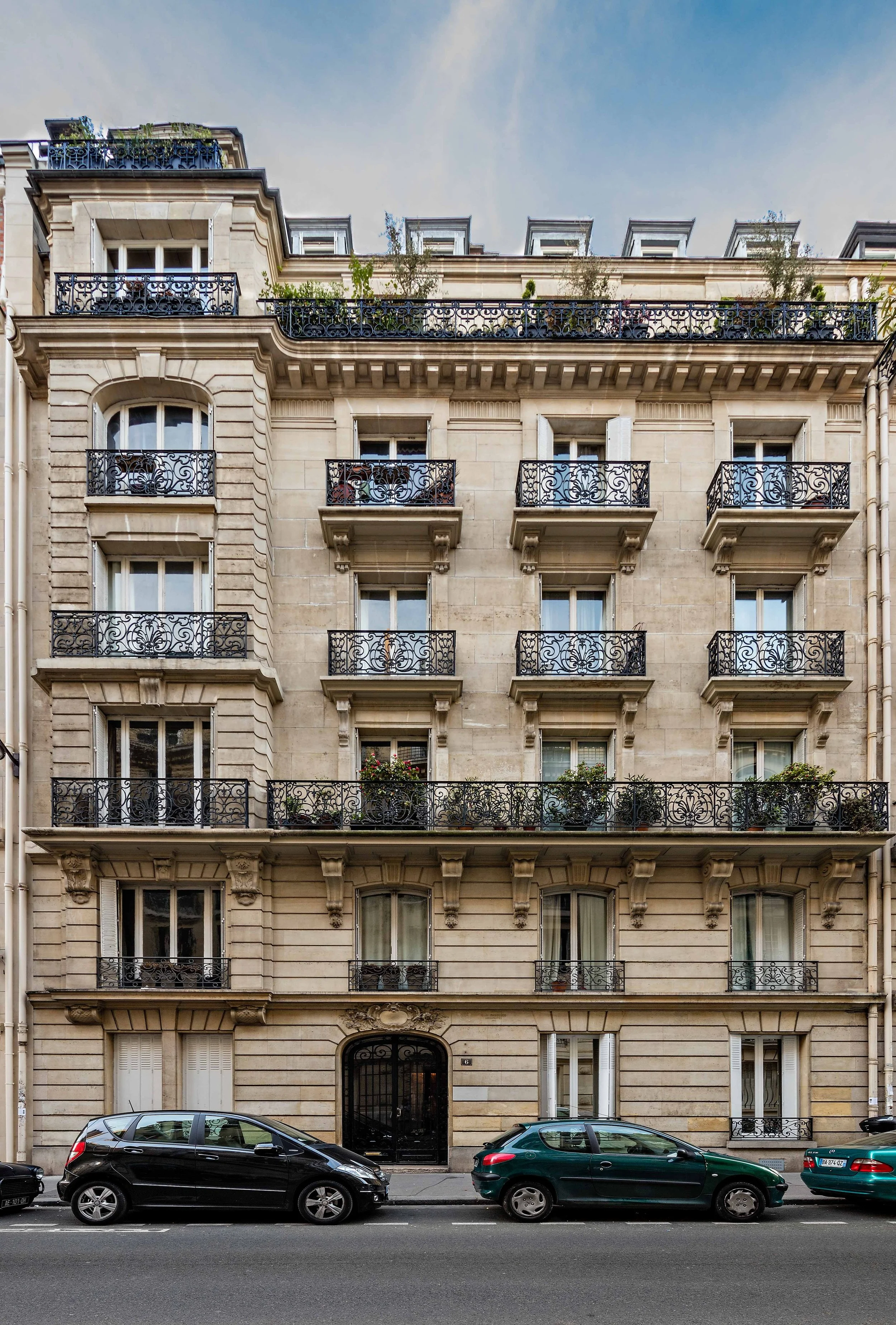 Immeuble haussmannien avec plusieurs étages, balcons en fer forgé, fenêtres à volets, et trois voitures garées devant.