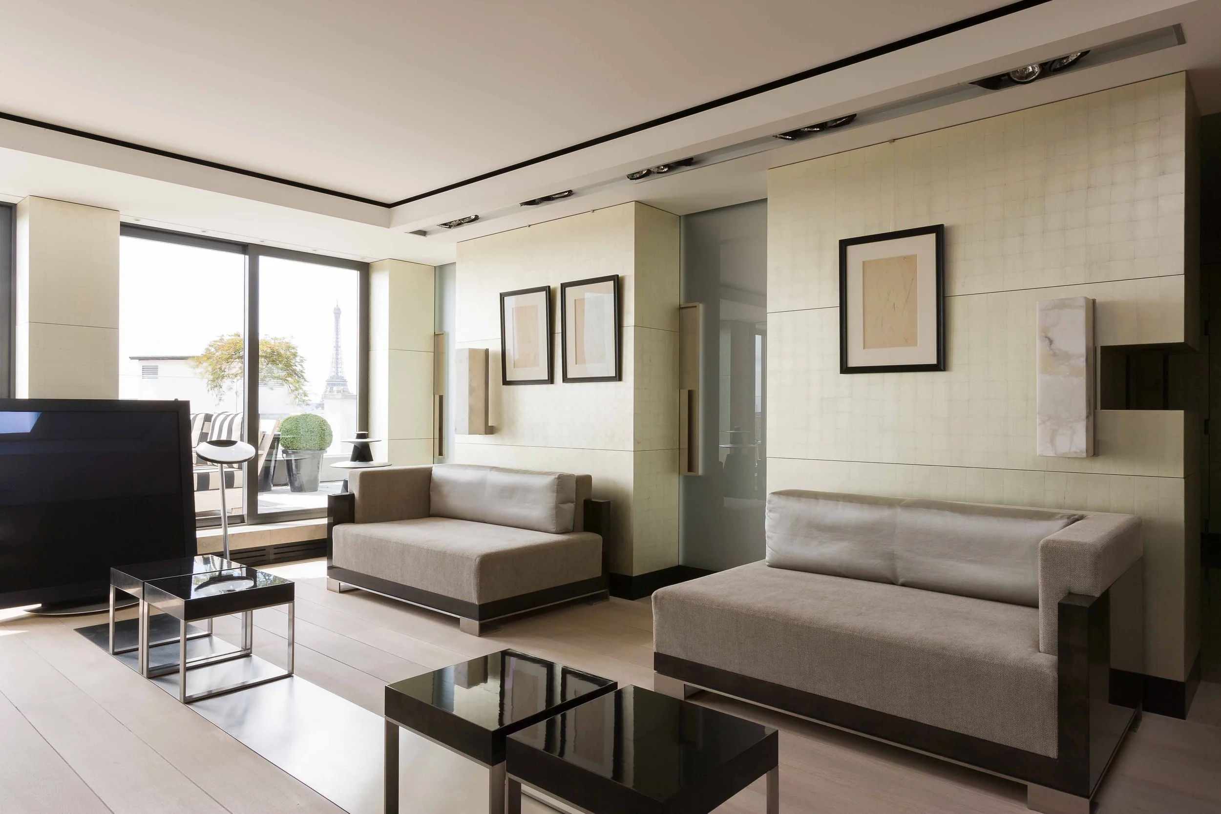 Salon contemporain avec deux canapés gris, tables basses en noir et argent, grande fenêtre avec vue sur la Tour Eiffel et un balcon avec des plantes.