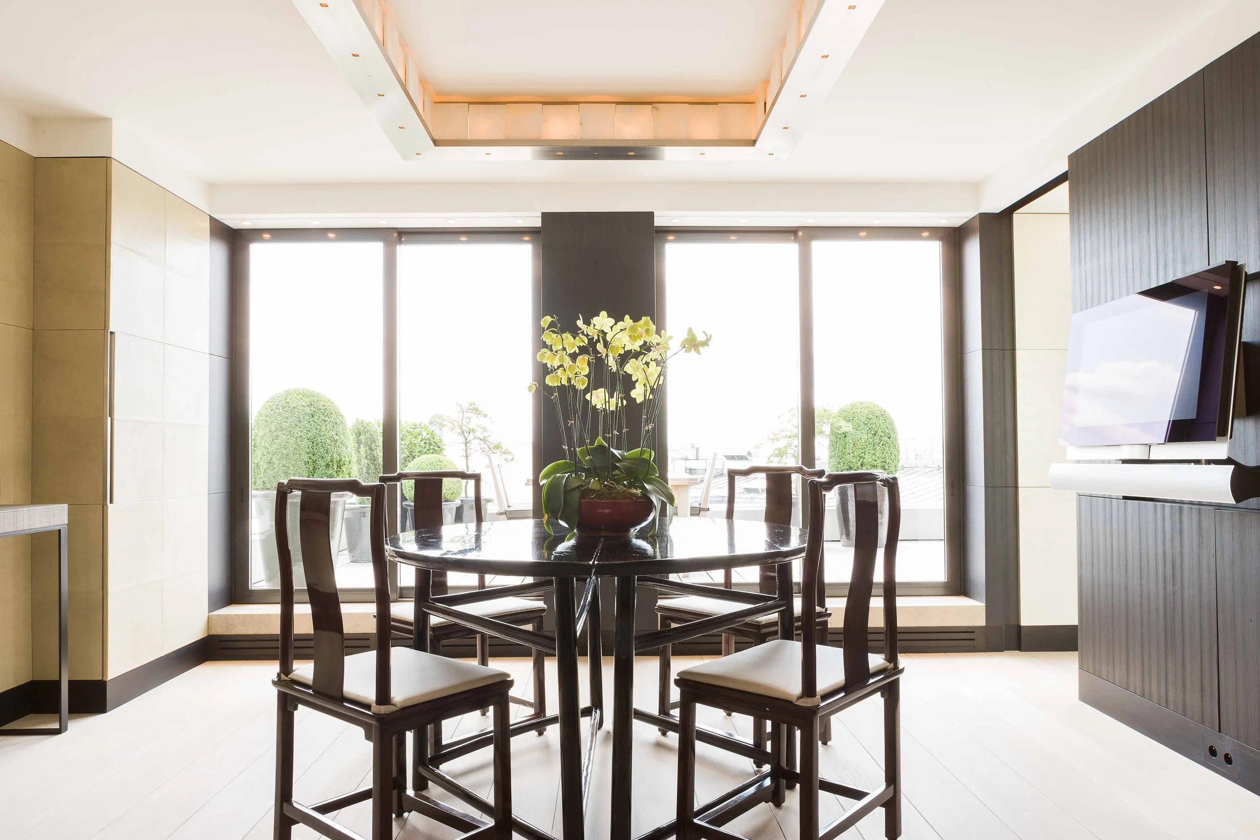 Salle à manger moderne avec table ronde, six chaises en bois, vase de fleurs blanches, grandes fenêtres avec vue sur des arbres et ciel en arrière-plan, télévision à écran plat sur le mur à droite.