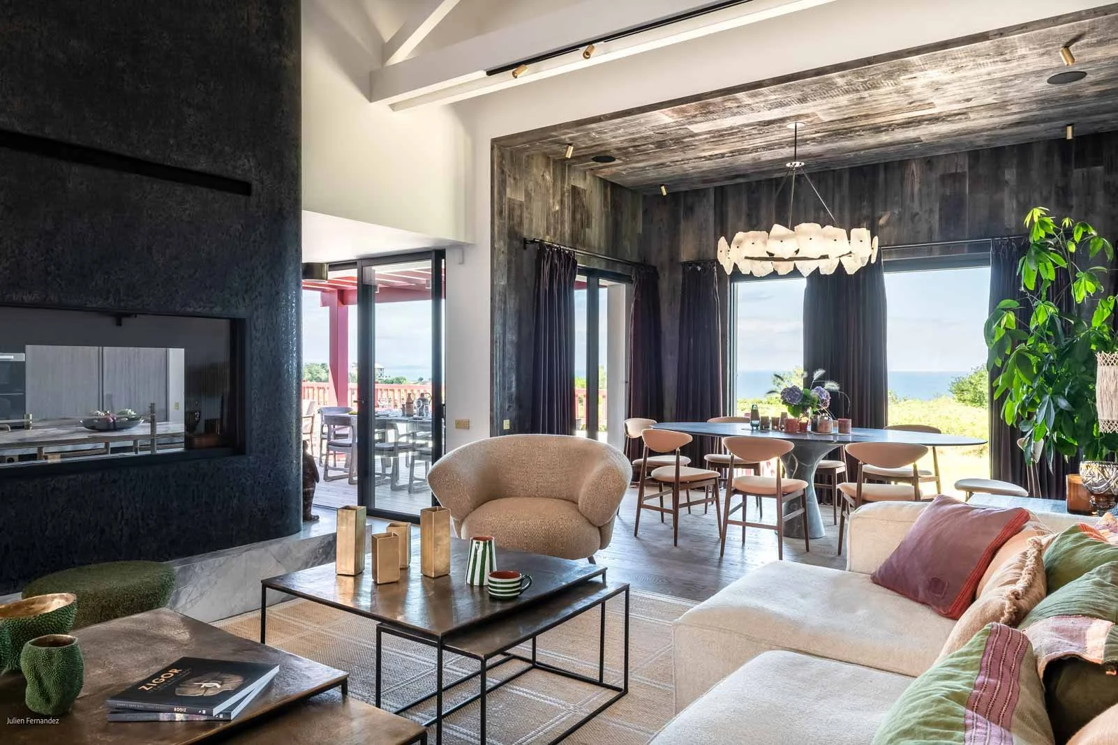 Salon lumineux avec un canapé beige, une table basse en métal, une cheminée noire, et une salle à manger avec vue sur la mer, décorée avec un chandelier blanc et des rideaux noirs.