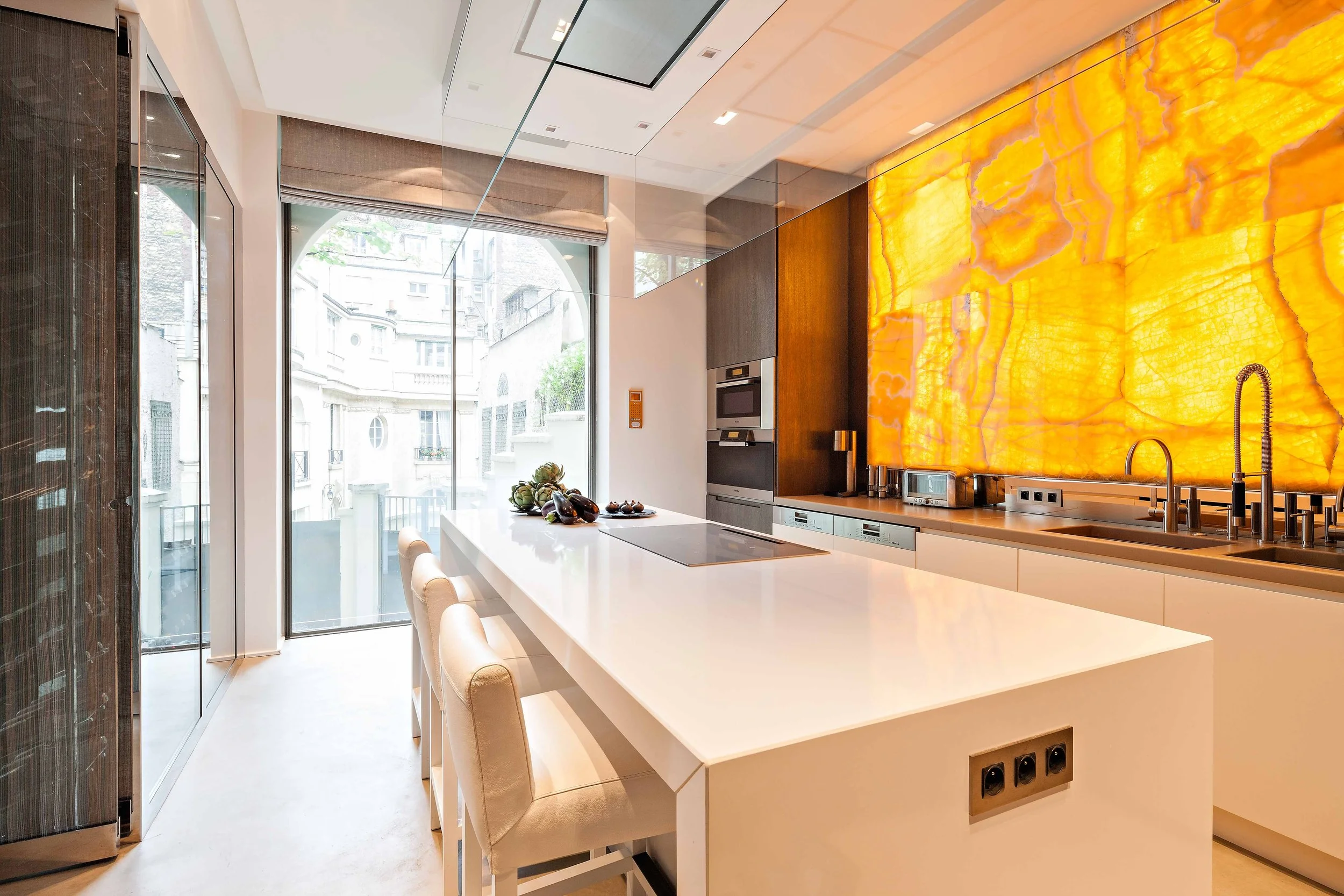 Cuisine moderne avec un grand îlot blanc, un mur en pierre de onyx éclairée, et une grande fenêtre laissant entrer la lumière naturelle.