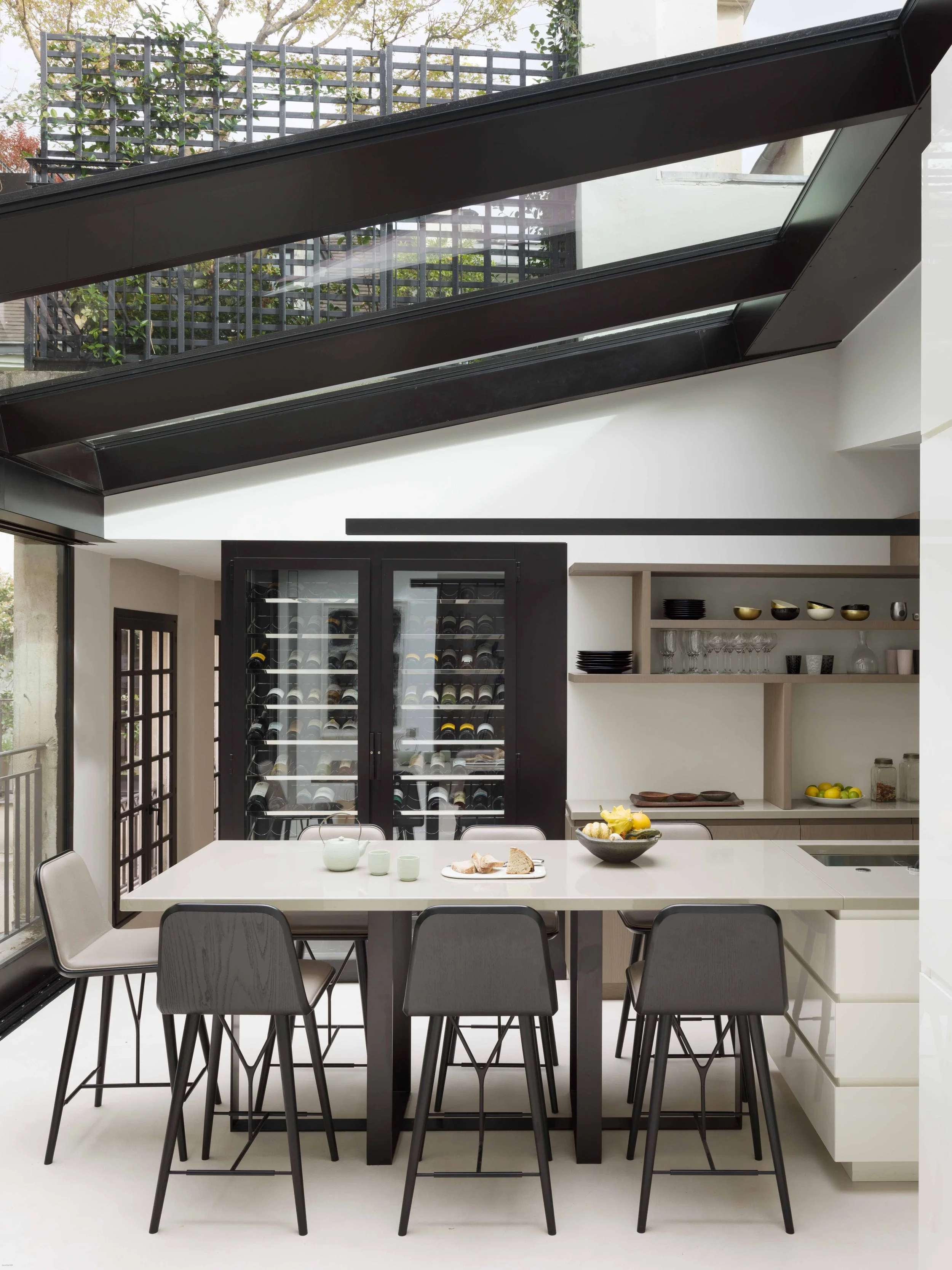 Cuisine moderne avec un grand îlot blanc, six chaises gris foncé, porte vitrée vers un cellier avec vin, étagères ouvertes avec vaisselle et verres, plafond en verre avec lumière naturelle.