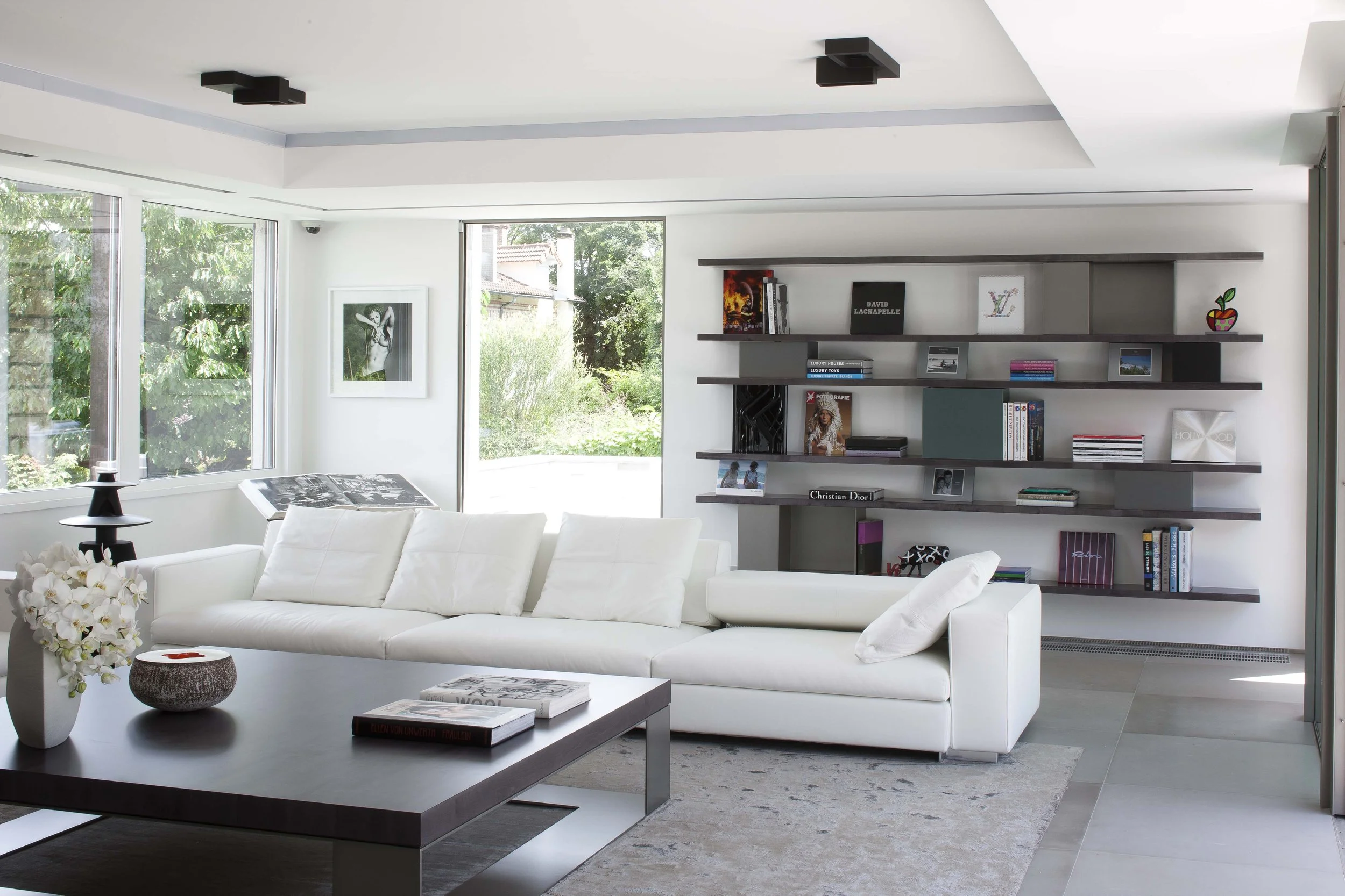 Salon moderne avec canapé blanc, table basse en bois foncé, étagères murales avec livres et objets décoratifs, grande fenêtre laissant entrer la lumière naturelle, vue sur un jardin extérieur.