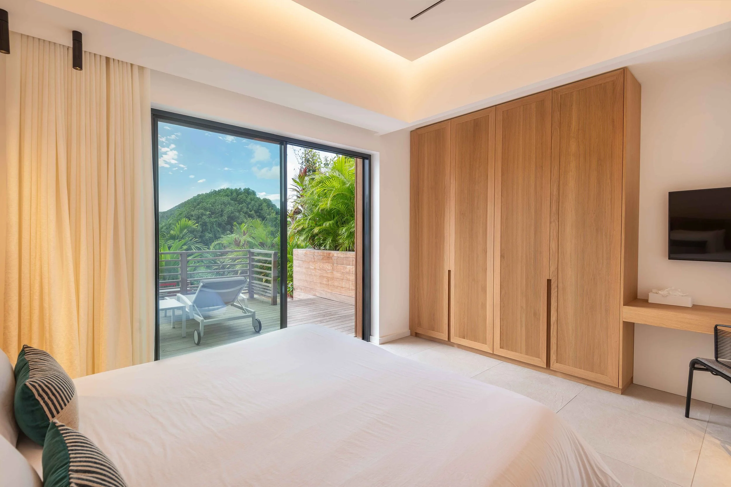 Chambre d'hôtel moderne avec lit blanc, coussins rayés, grands miroirs, vue sur un balcon avec fauteuils en bois et vue sur la nature, porte-fenêtre coulissante, mobilier en bois clair, télévision à écran plat, ambiance chaleureuse et lumineuse.