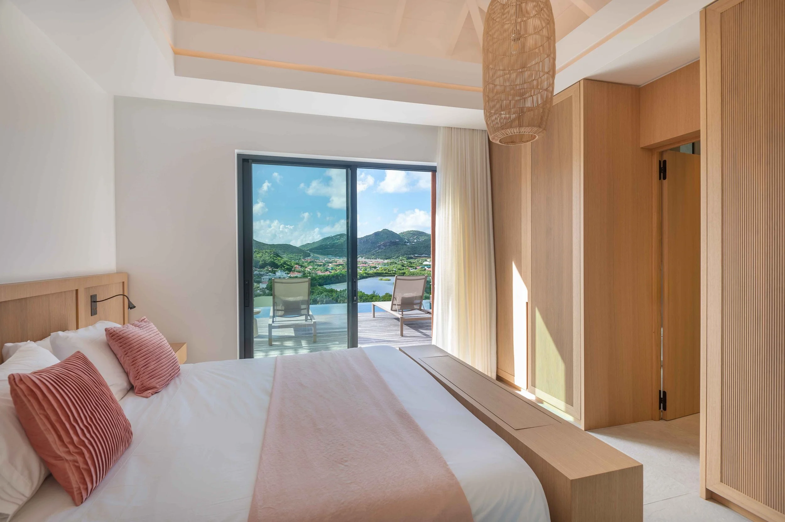 Chambre avec lit à linge blanc, oreillers roses, vue sur un balcon avec fauteuils et un paysage de montagnes et de rivière sous un ciel bleu.
