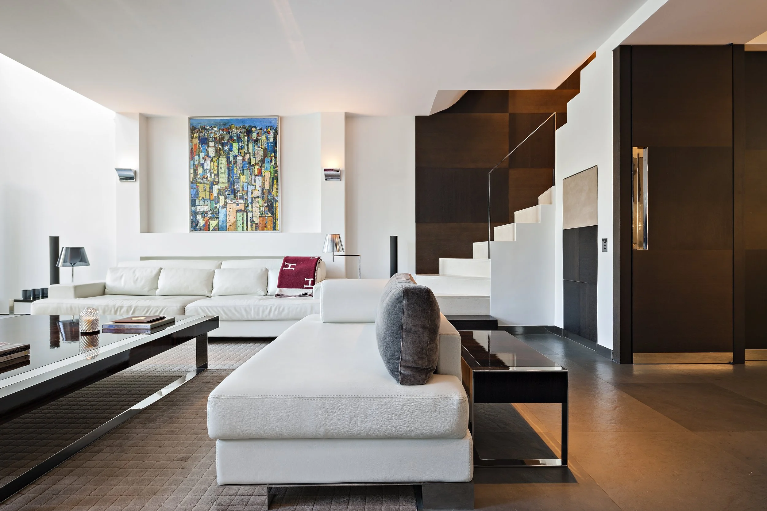 Salon moderne avec canapé blanc, tableau coloré au mur, escalier en bois blanc, murs blancs et meubles en bois foncé.