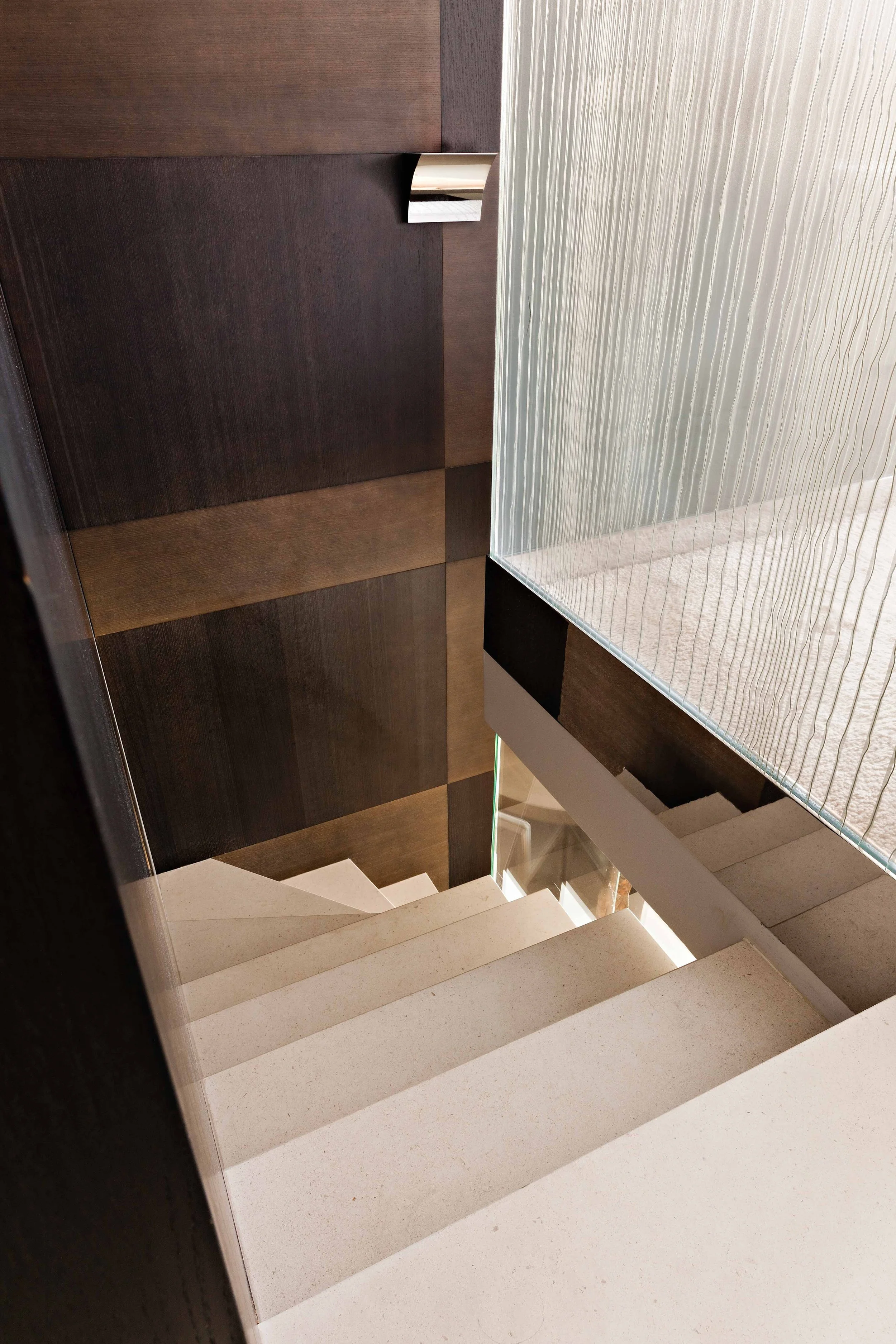 Vue en plongée d'un escalier intérieur moderne avec marches en pierre blanche, mur en bois foncé et panneau en verre ondulé.