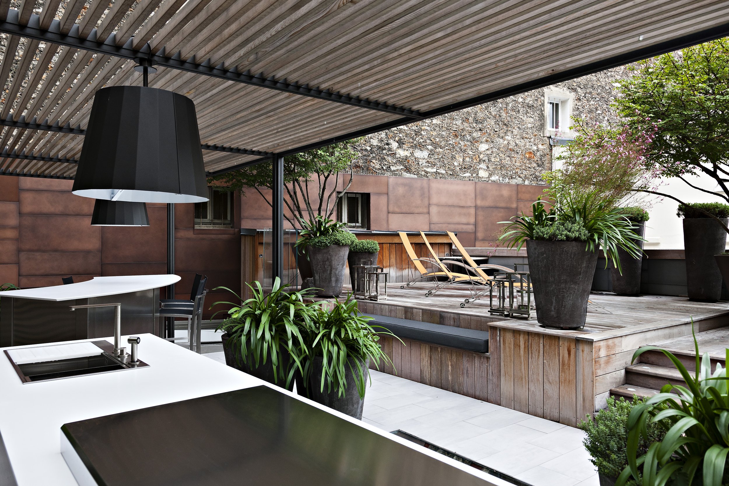 Une terrasse extérieure avec des plantes en pot, des chaises longues en bois, et un toit en lamelles de bois.