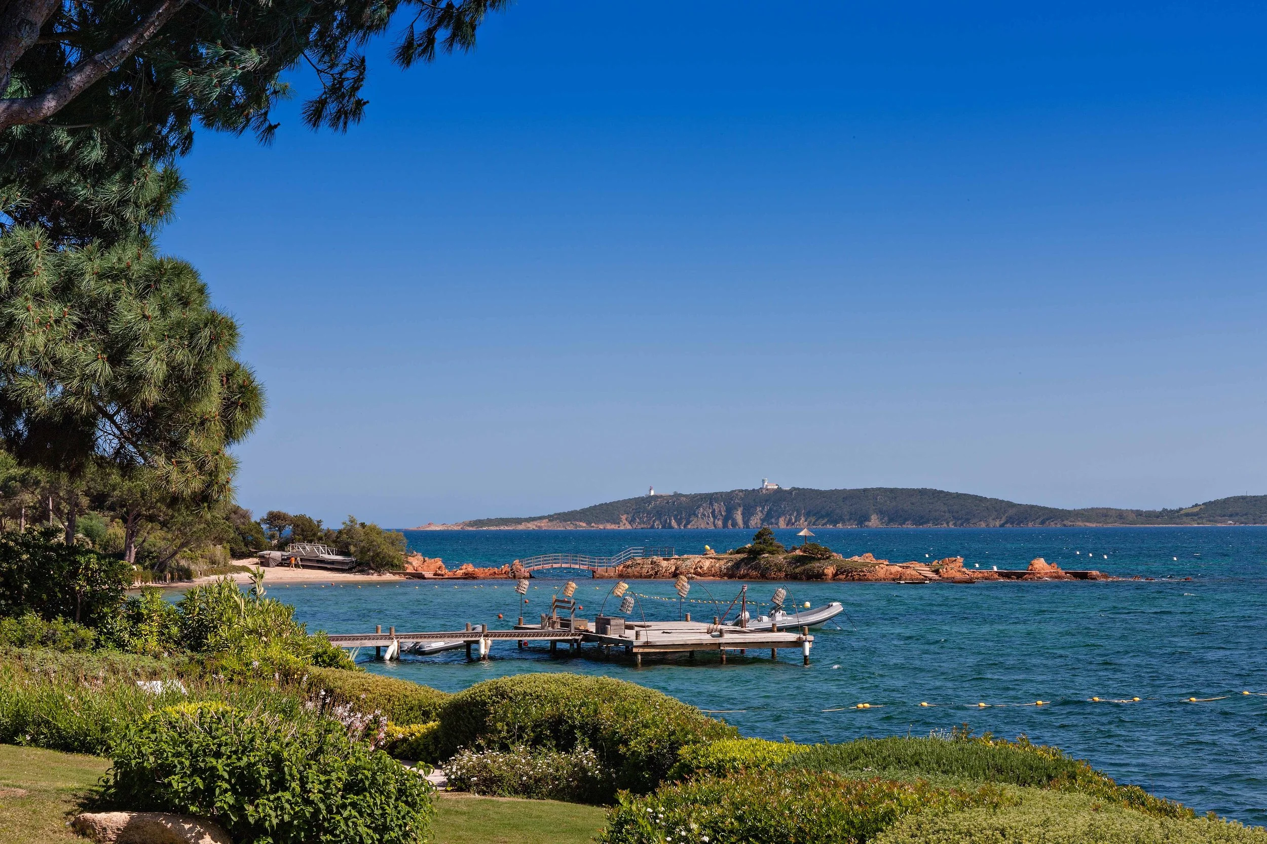 Paysage côtier avec un ponton, mer bleue, rochers et île au loin, arbres et buissons verts au premier plan, ciel clair.