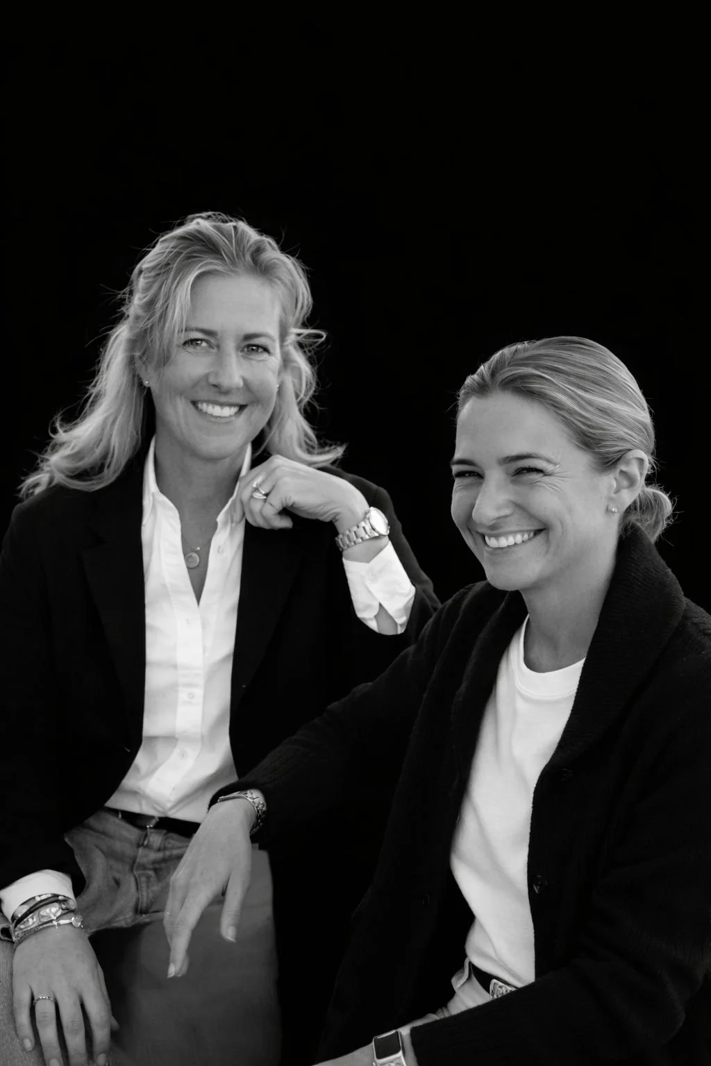 Deux femmes souriantes posant ensemble, l'une en blazer et chemise blanche, l'autre en sweat à capuche noire, en noir et blanc.