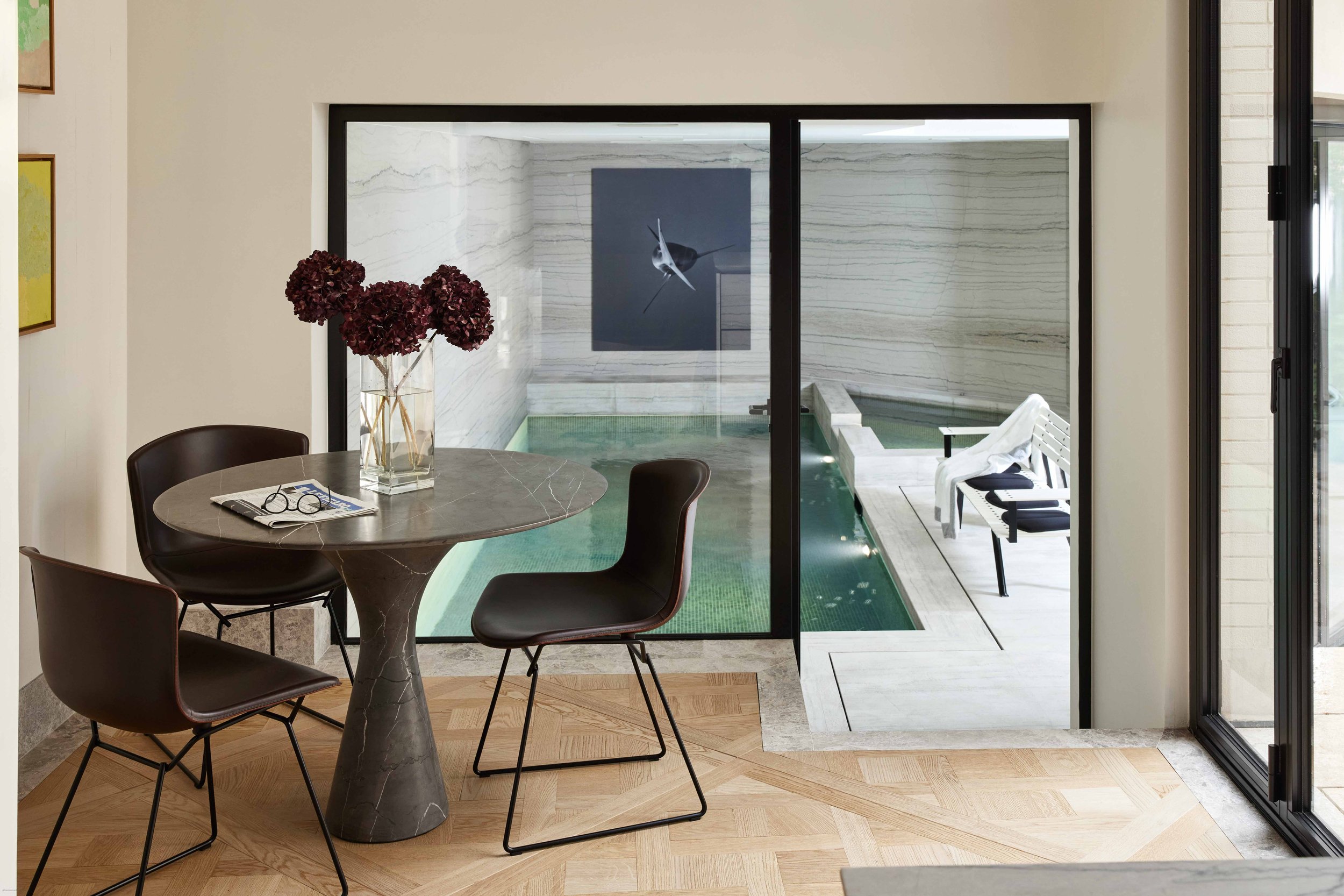 Salle à manger intérieure avec table en marbre, trois chaises en cuir, vase de fleurs violettes, vue sur un petit bassin d'eau à travers une grande fenêtre en verre.