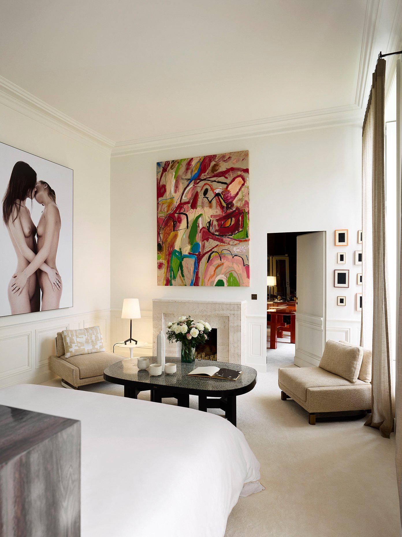 Salon de chambre avec mur blanc, grand tableau abstrait coloré, grande fenêtre avec rideaux, canapé beige, table basse ronde avec livre et fleurs, deux petits fauteuils, cheminée, lampe de chevet.