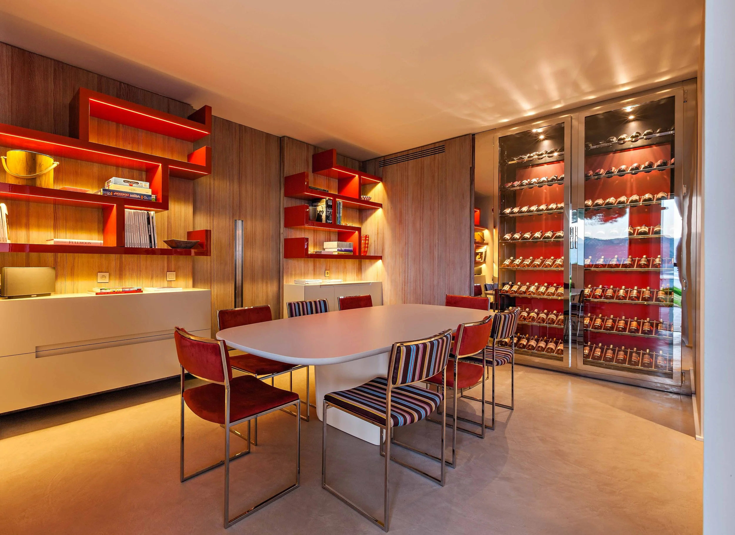 Salle de réunion avec table ovale, chaises rembourrées et un grand réfrigérateur à vin en verre rempli de bouteilles. Murs en bois, étagères rouges avec livres et décorations.