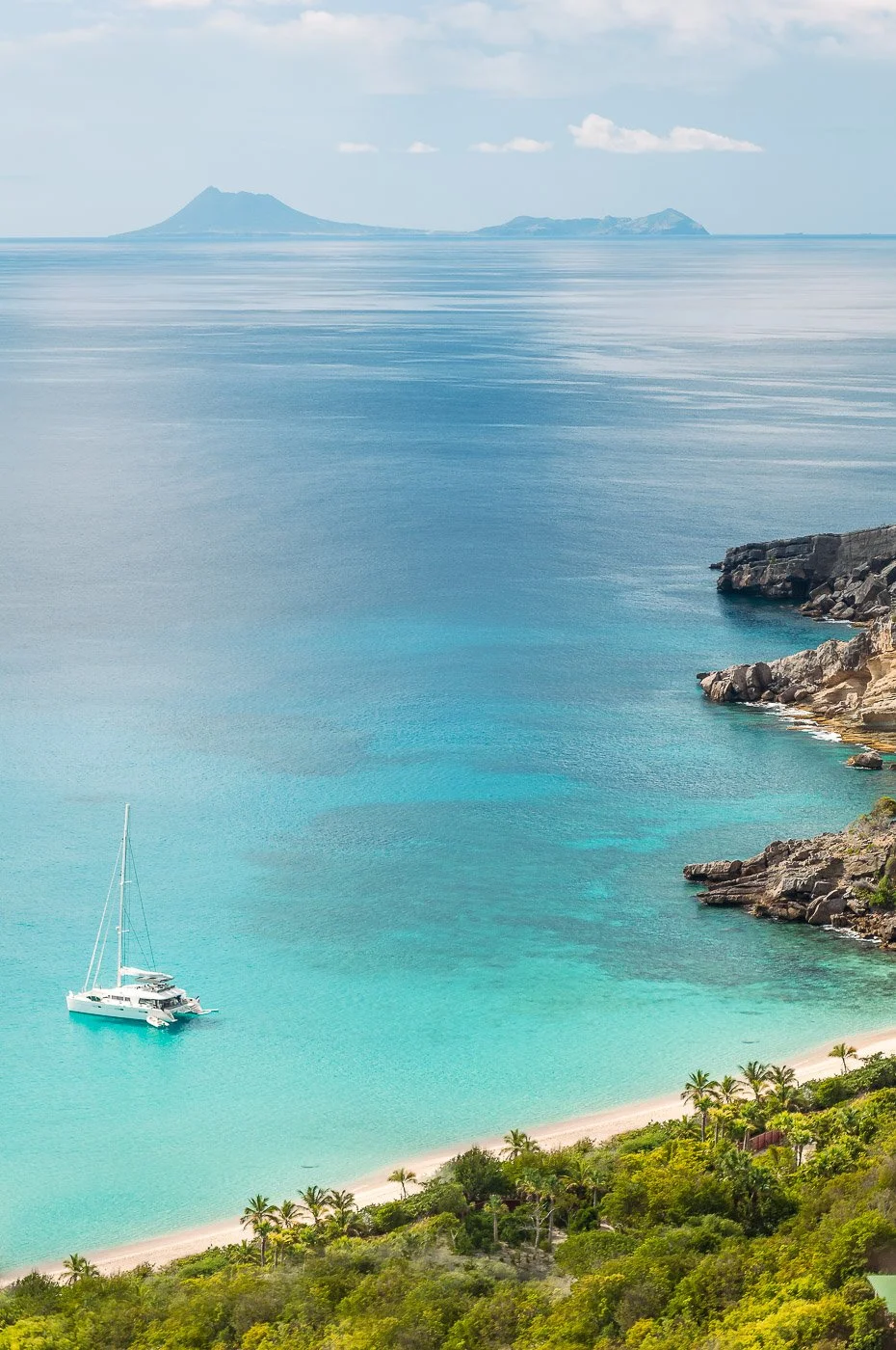 Une plage tropicale avec des eaux turquoise, un voilier au mouillage, une côte rocheuse et une végétation luxuriante, avec une île en arrière-plan.