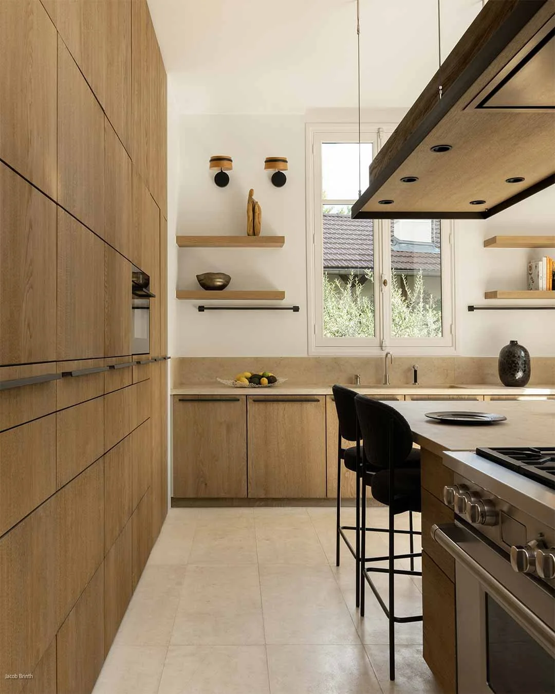 Cuisine moderne avec armoires en bois clair, comptoir en pierre beige, fenêtres laissant entrer la lumière naturelle, étagères en bois avec des décorations, table avec deux chaises noires.