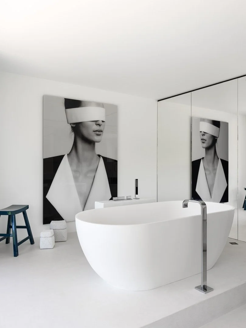 Salle de bain moderne avec une baignoire blanche ovale, deux grands tableaux en noir et blanc représentant des femmes avec un bandeau blanc sur les yeux, un petit tabouret bleu, deux petits cubes blancs et un miroir mural.