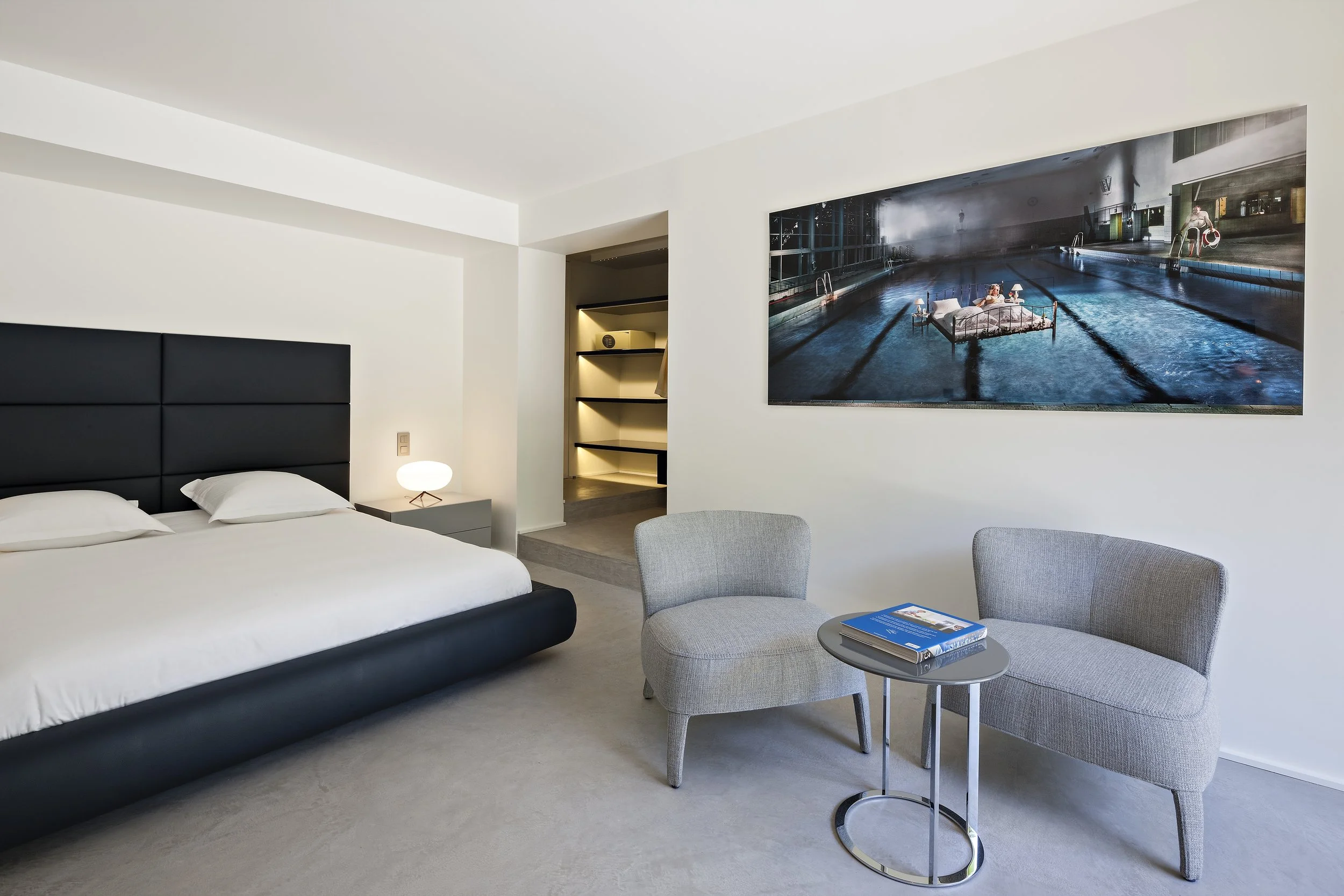 Chambre d'hôtel moderne avec lit king-size, deux fauteuils gris, petite table ronde avec des livres, grande photo d'une piscine intérieure sur le mur, et un éclairage doux.