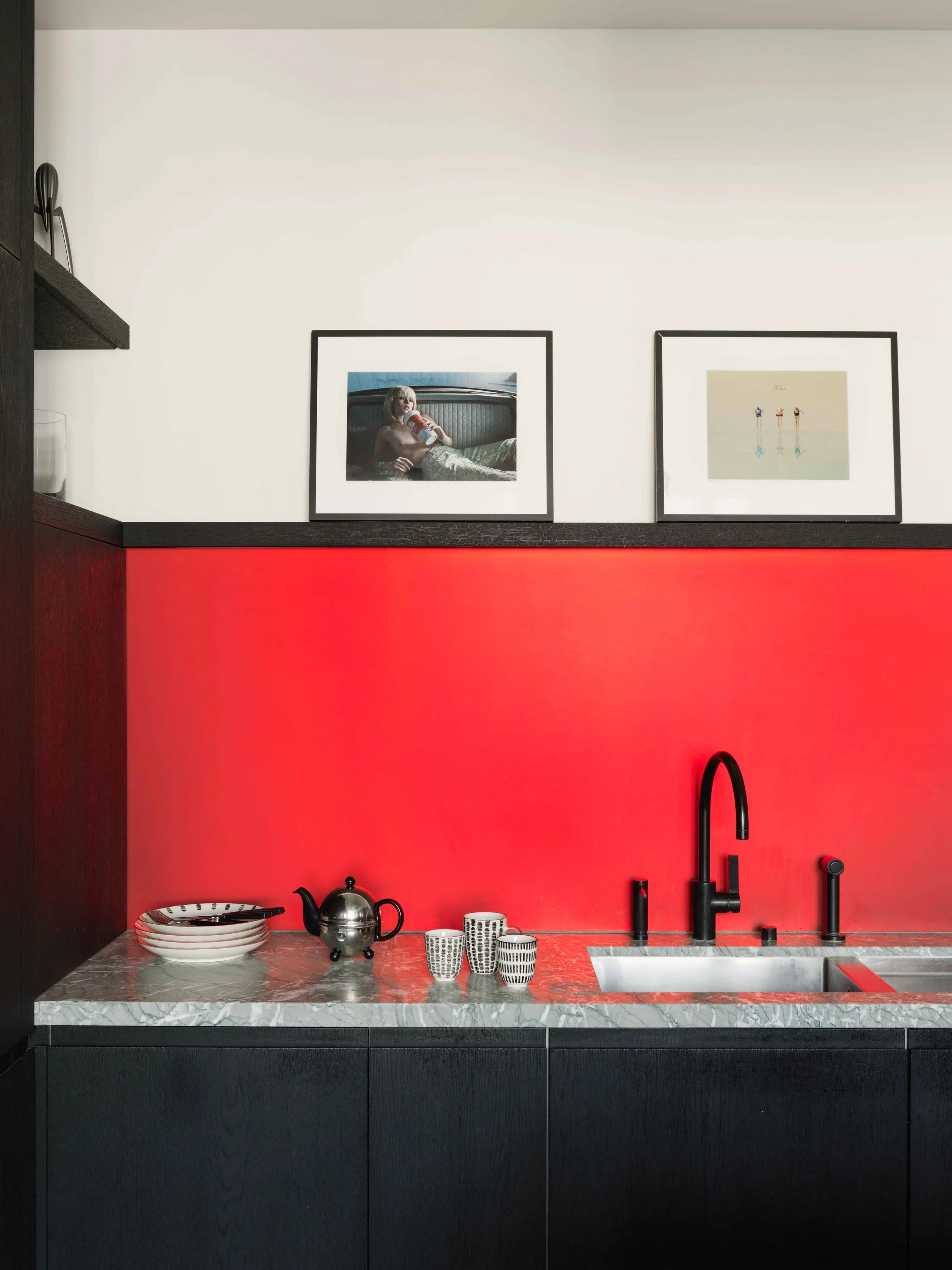 Cuisine moderne avec un plan de travail en marbre, un évier noir, des tasses et une théière en métal, un mur rouge en arrière-plan et deux cadres photo noirs au mur.
