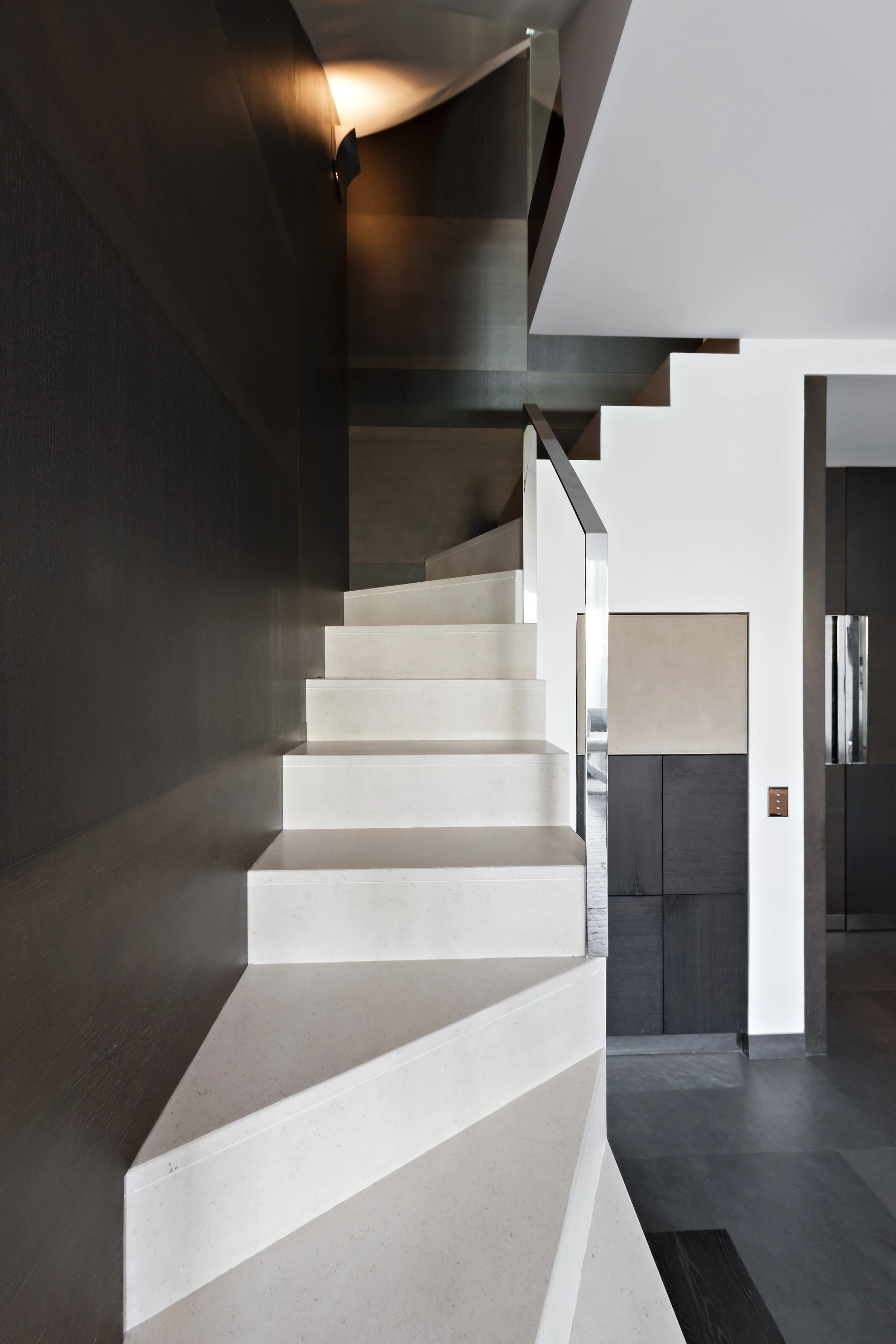 Escalier moderne avec marches blanches, mur en bois foncé à gauche, murs blancs et un interrupteur dans une pièce contemporaine.
