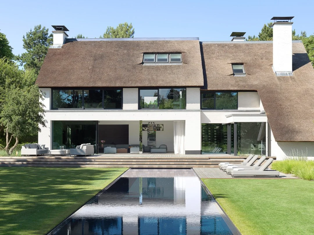 Maison moderne avec toit en chaume, grande terrasse, piscine rectangulaire et mobilier extérieur, entourée d'arbres et d'un jardin bien entretenu.