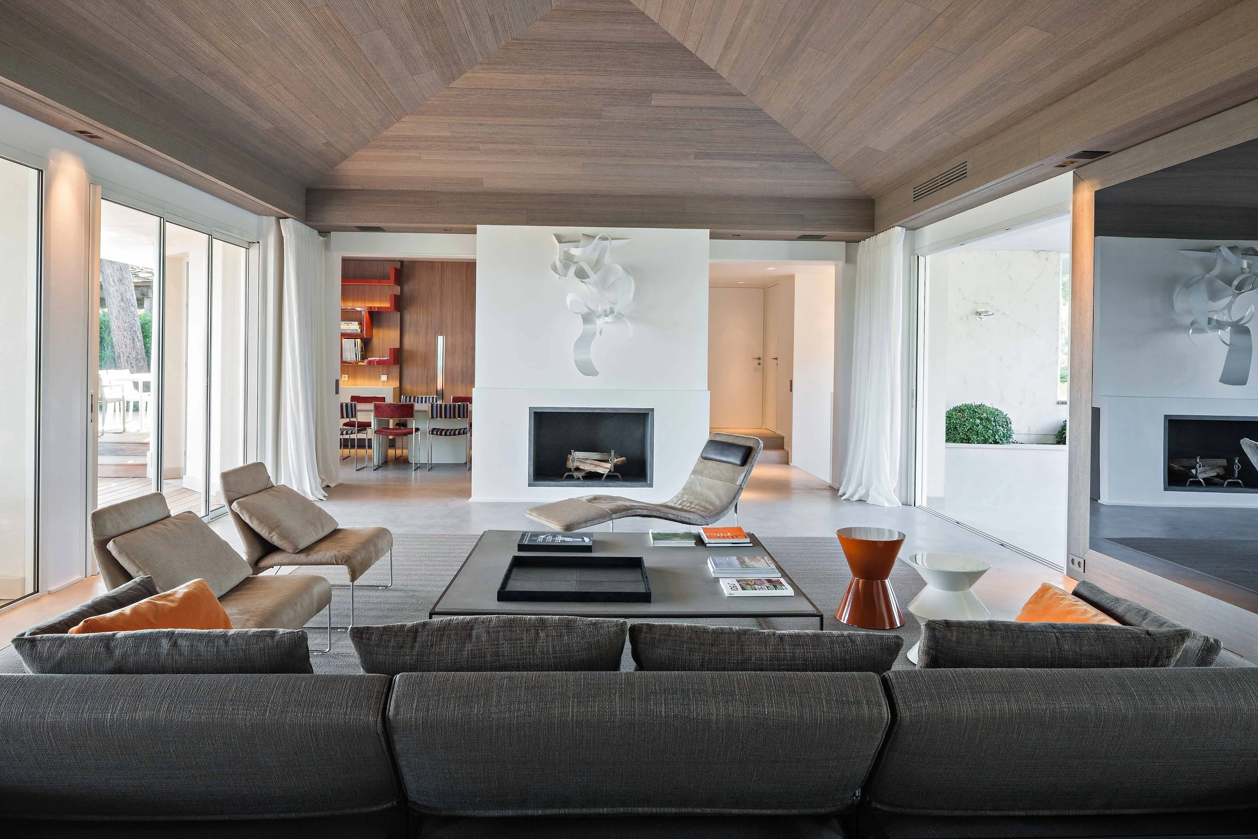 Salon moderne avec canapé gris, fauteuils en beige, cheminée blanche, décoration artistique murale blanche, grande pièce lumineuse avec vue sur l'extérieur