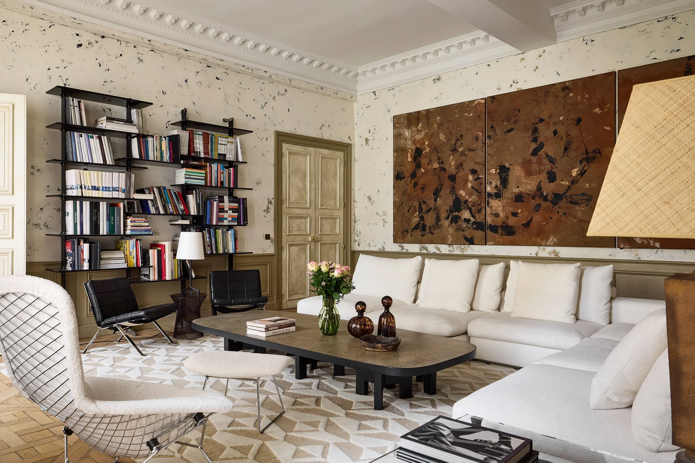 Salon moderne avec canapé blanc en forme de L, table basse, fauteuils noirs, bibliothèque noirs pleine de livres, mur avec peinture abstraite, lampe en bois, vase avec fleurs, tapis beige et blanc, mur en pierre beige, plafond orné de moulures.