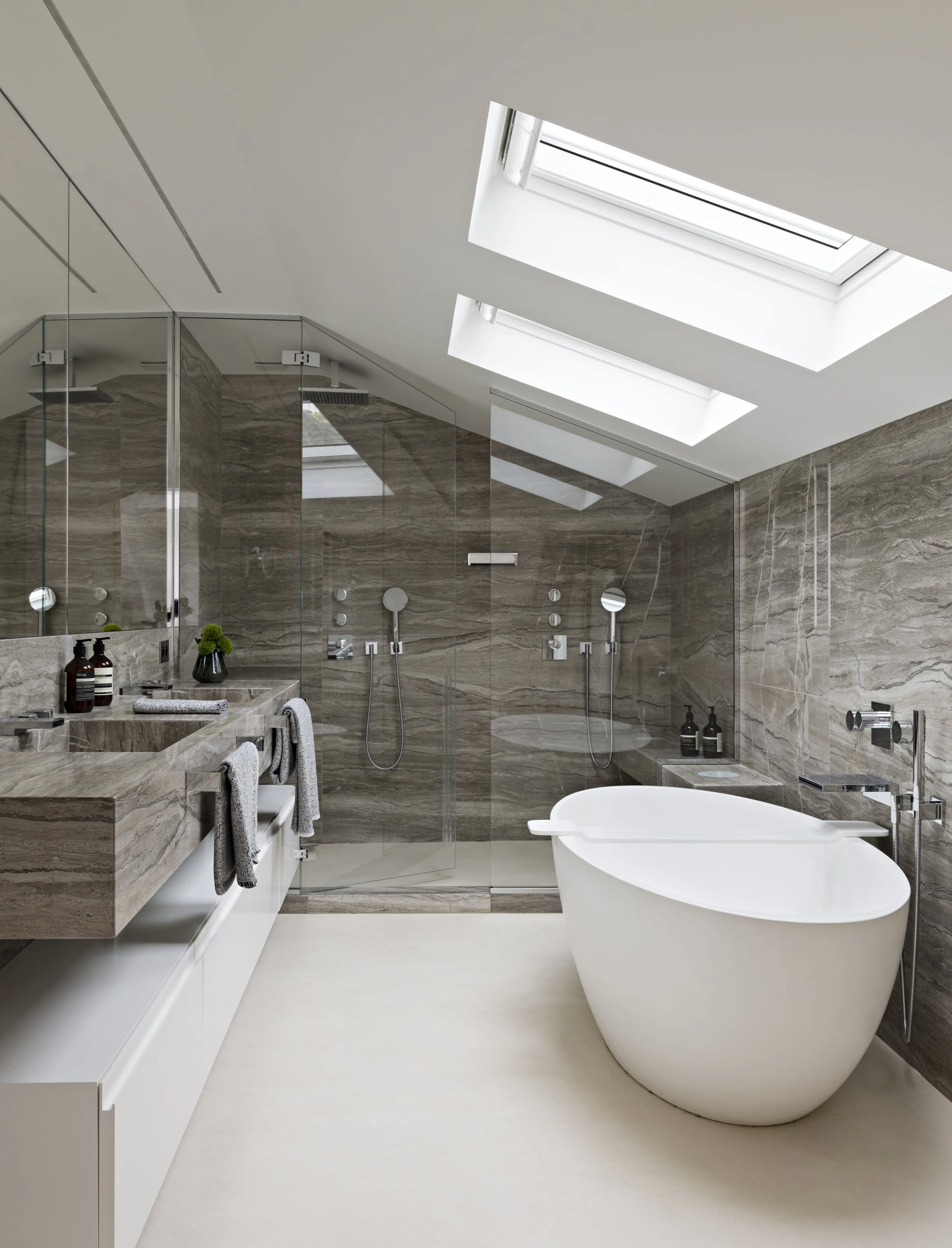 Salle de bain moderne avec douche à l'italienne, baignoire autoportante blanche, murs recouverts de marbre, grande fenêtre de toit, lavabo en marbre, serviettes suspendues, produits de bain sur un petit meuble.
