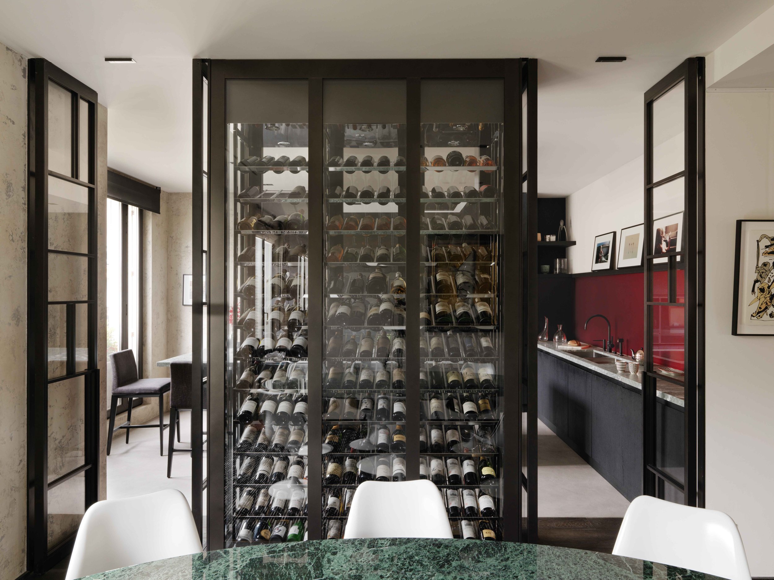 Une cave à vin moderne avec un mur en verre contenant de nombreuses bouteilles de vin, un coin repas avec un tableau et une table en marbre vert, un évier dans un espace de cuisine en arrière-plan.