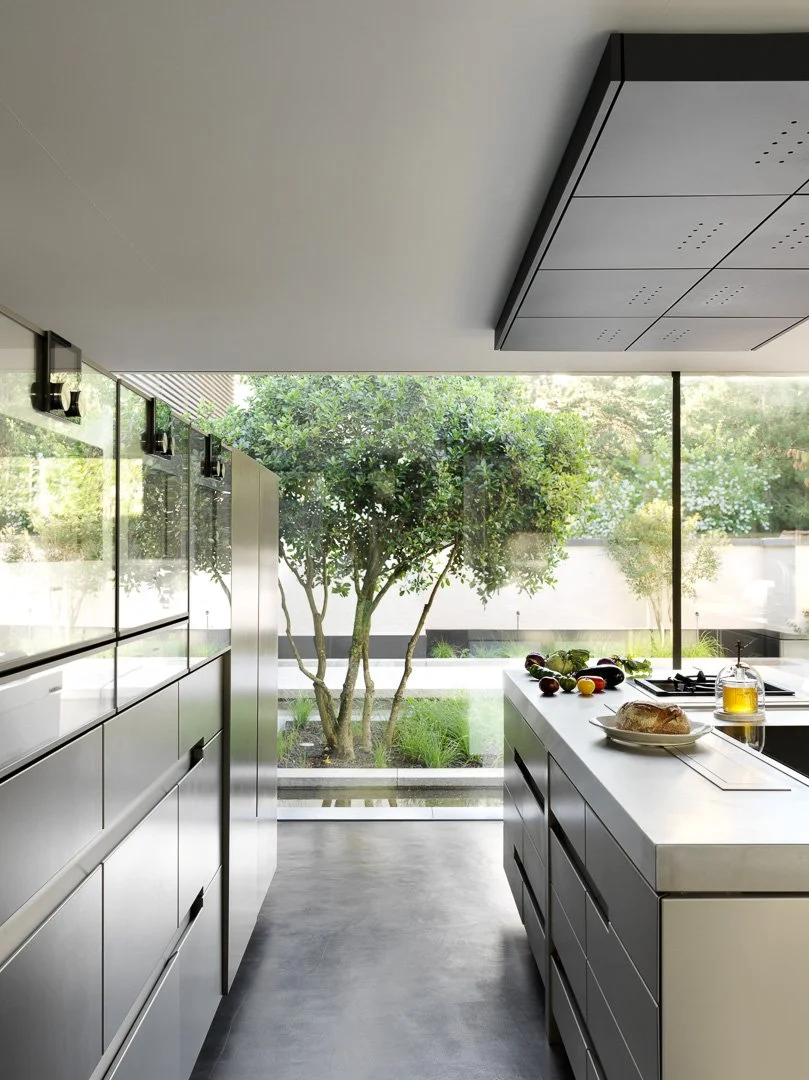 Cuisine moderne avec grand mur de fenêtres donnant sur un jardin, plan de travail blanc avec des fruits, un pain et de l'huile.