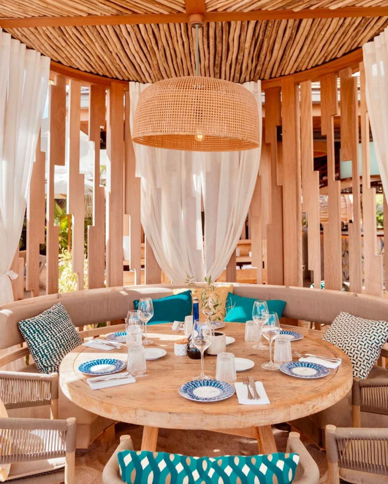 Table ronde en bois avec assiettes, verres à vin, ustensiles, serviettes, et coussins bleus et noirs, dans un espace en bois avec rideaux blancs et éclairage suspendu en rotin.