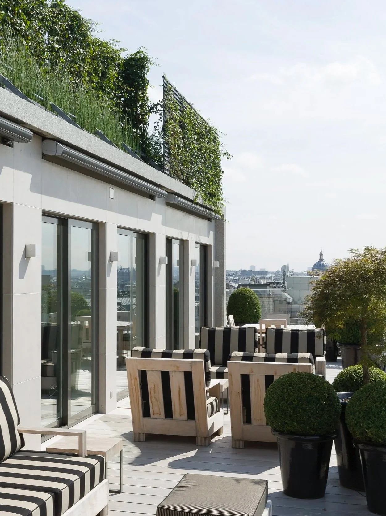 Terrasse urbaine avec mobilier en bois et coussins rayés noir et blanc, entourée de plantes en pot, avec vue sur la ville et le ciel en arrière-plan.
