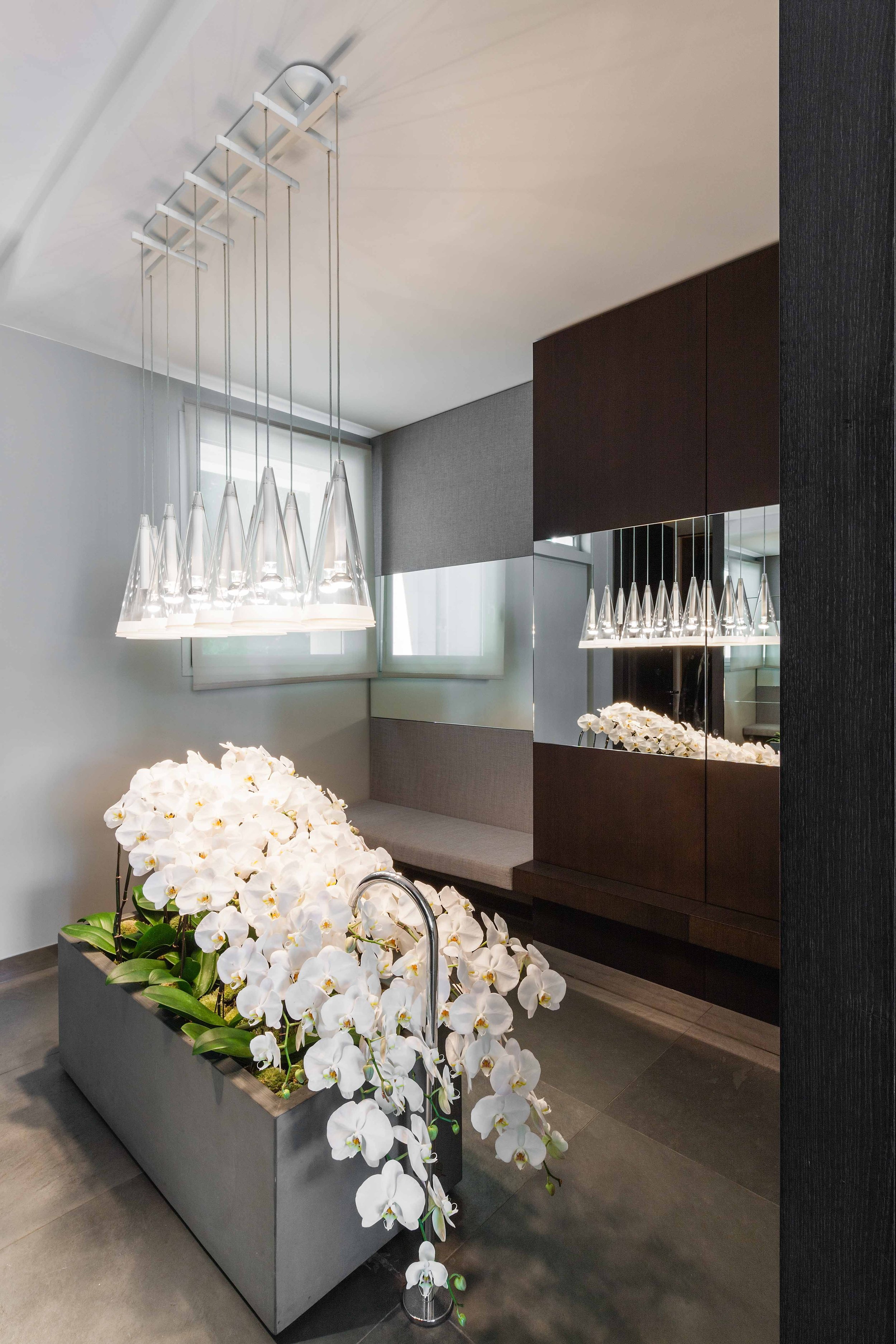 Vase rectangulaire avec des orchidées blanches dans une pièce moderne avec un luminaire design suspendu et un mur avec miroir.