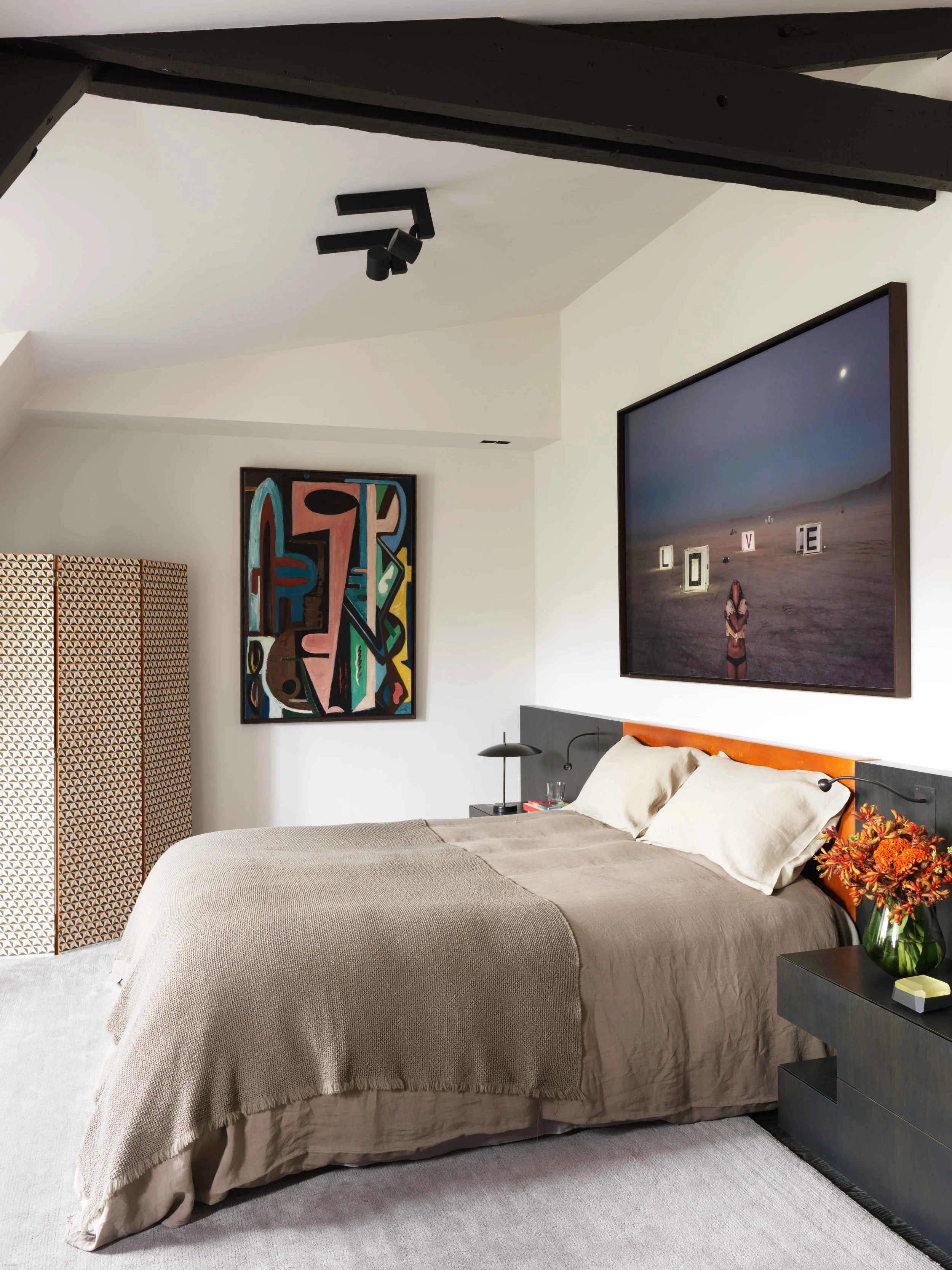 Chambre moderne avec un lit, deux oreillers, une lampe de chevet, une grande peinture abstraite sur le mur, une grande télévision affichant le mot 'LOVE' sur un fond de désert, et un vase avec des fleurs sur une table de nuit.