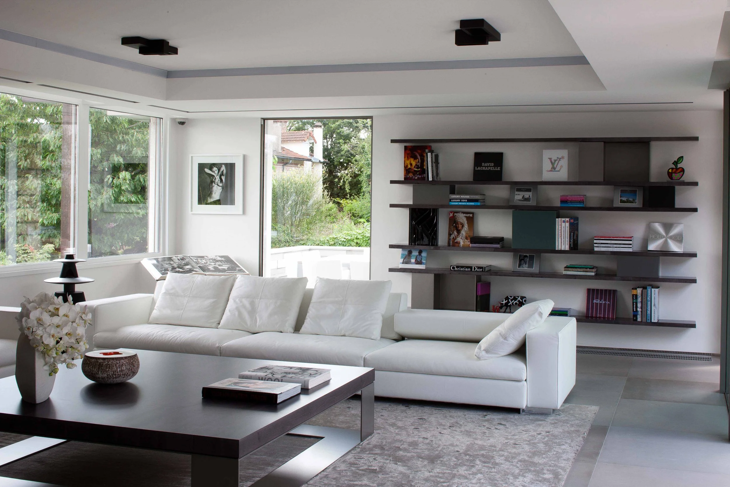 Salon moderne avec canapé blanc, table basse, étagères murales avec livres et décorations, grande fenêtre laissant entrer la lumière naturelle, tableau noir et blanc suspendu